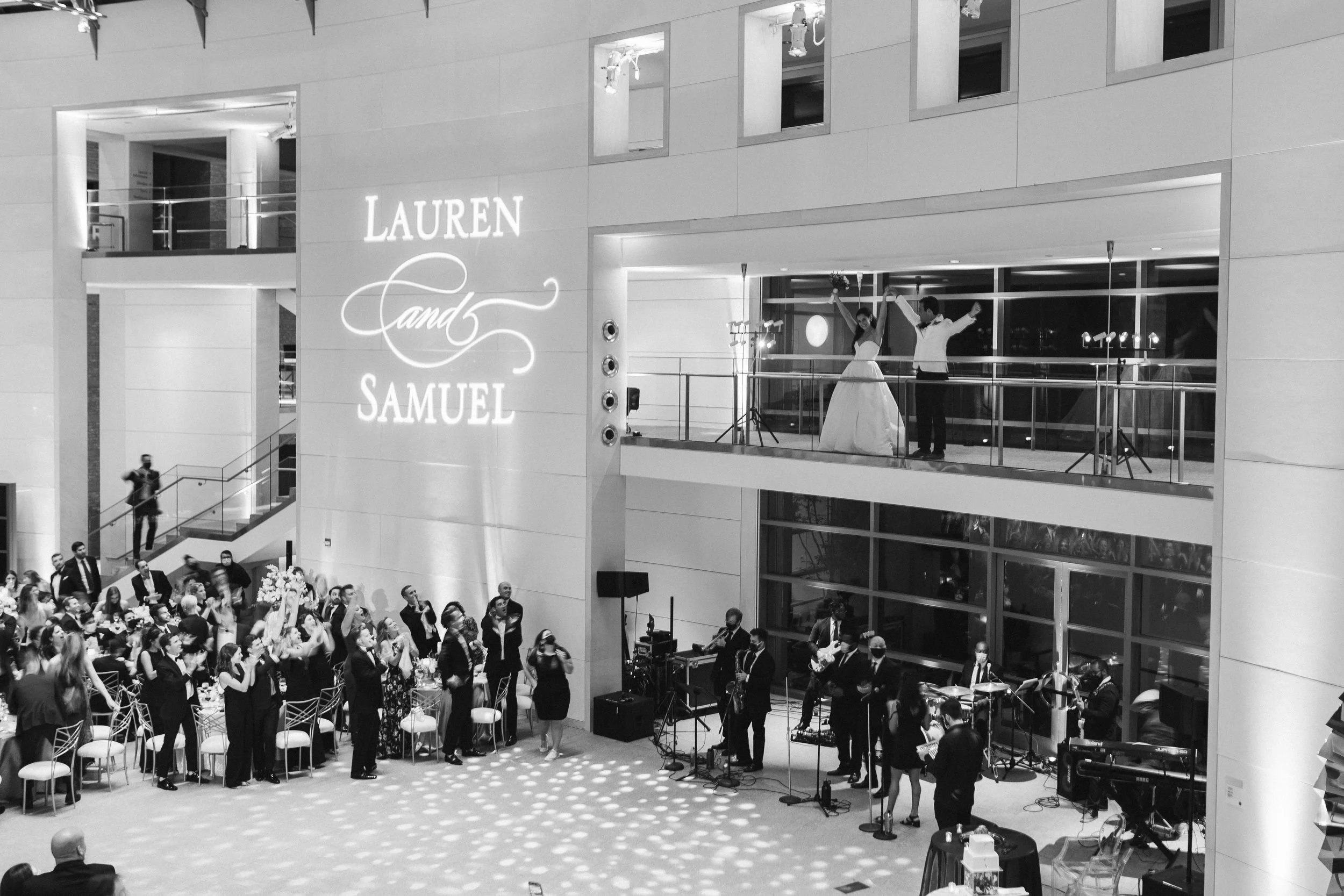 A black and white photo of a wedding reception inside a large modern building. The bride and groom are dancing on a balcony stage, holding hands and raising their arms. The crowd below is standing and facing the stage, with some people seated at tabl