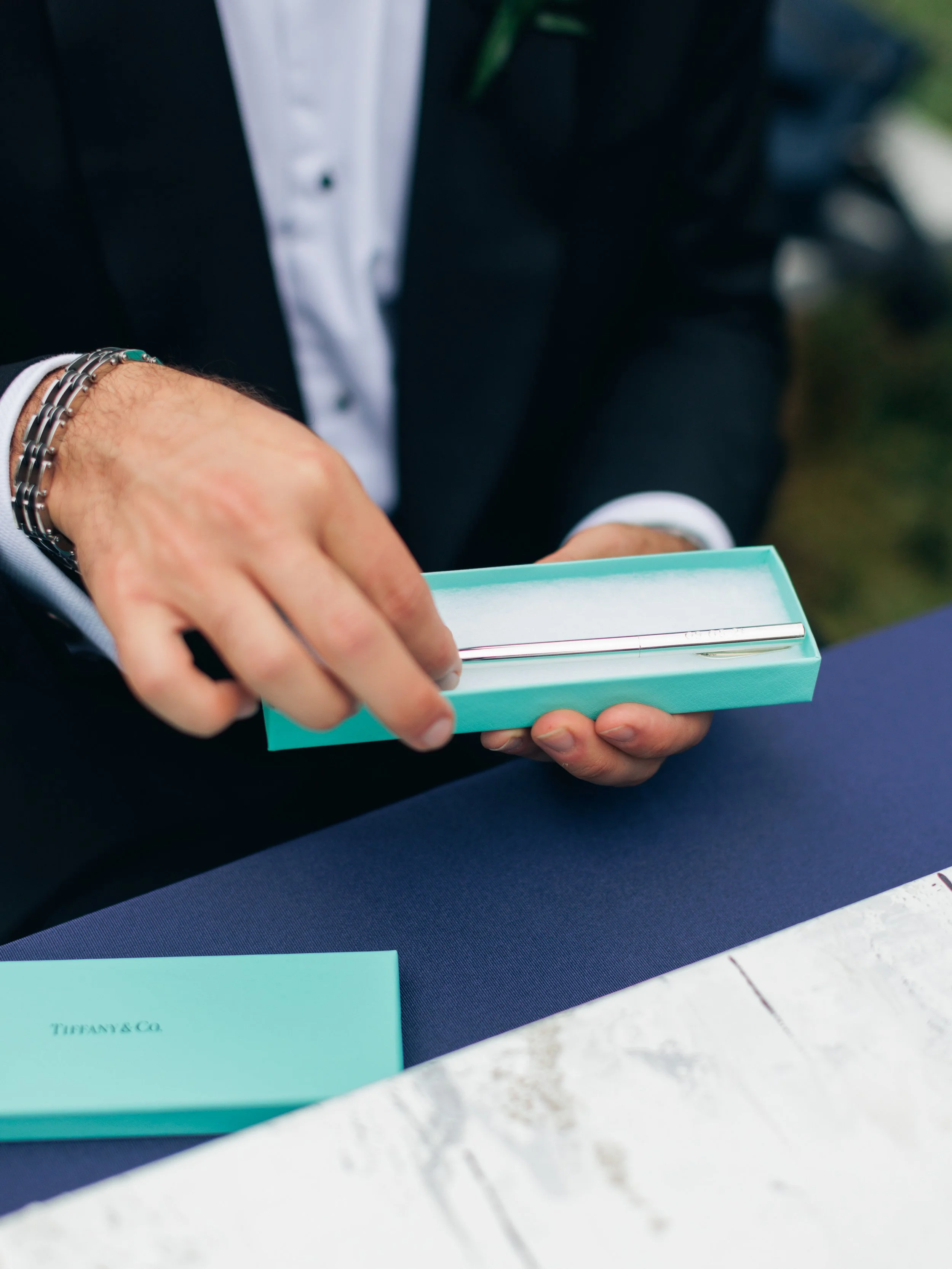 Close-up of a person's hand holding a Tiffany & Co. box, with a partial view of a tuxedo, including a white shirt and black jacket, in the background.