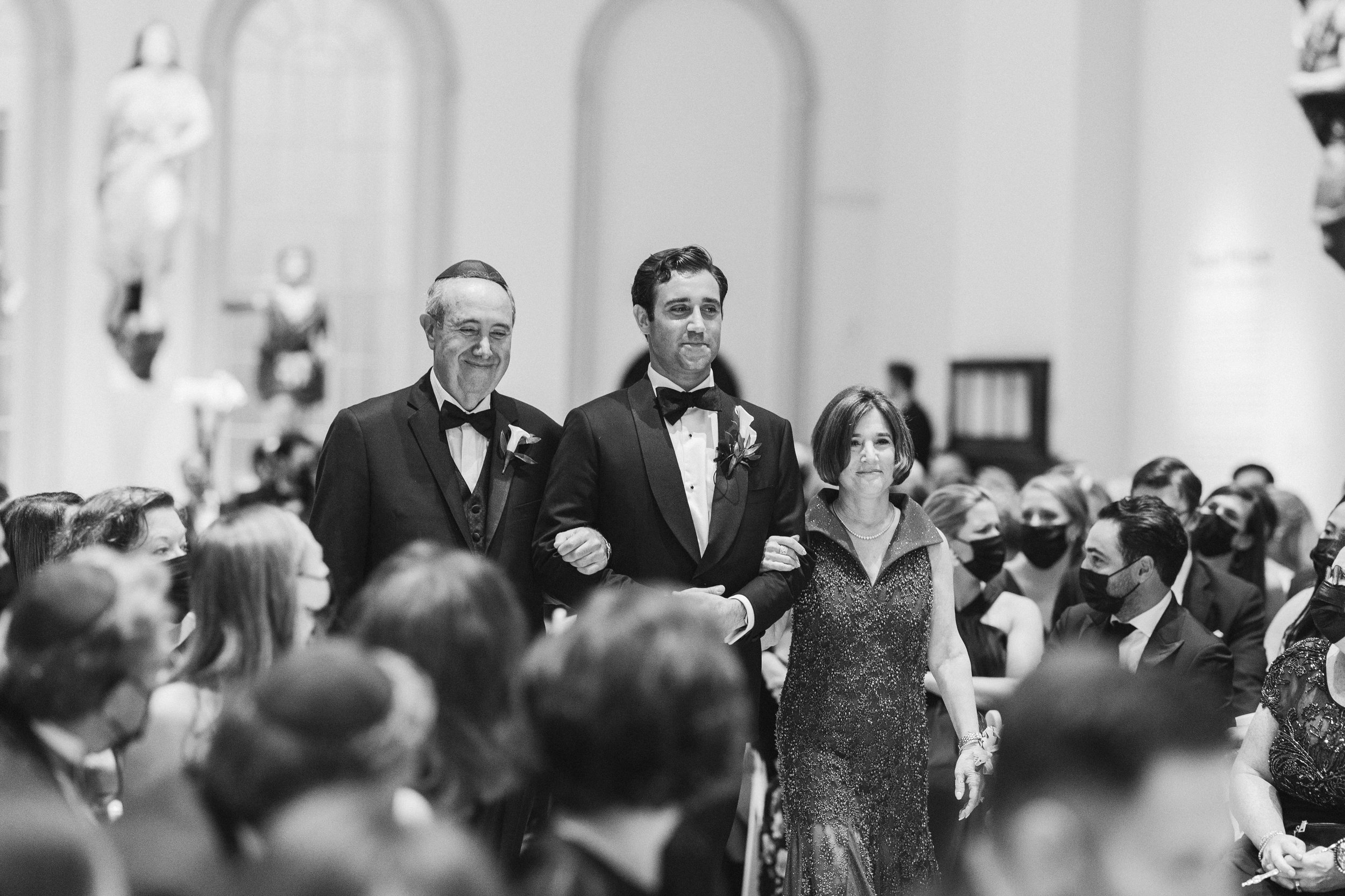 A black and white photo of a formal event with three people walking down an aisle. The man in the center is wearing a tuxedo with a bow tie, and an older man to his left is also dressed formally with a boutonniere. The woman on his right is in a spar