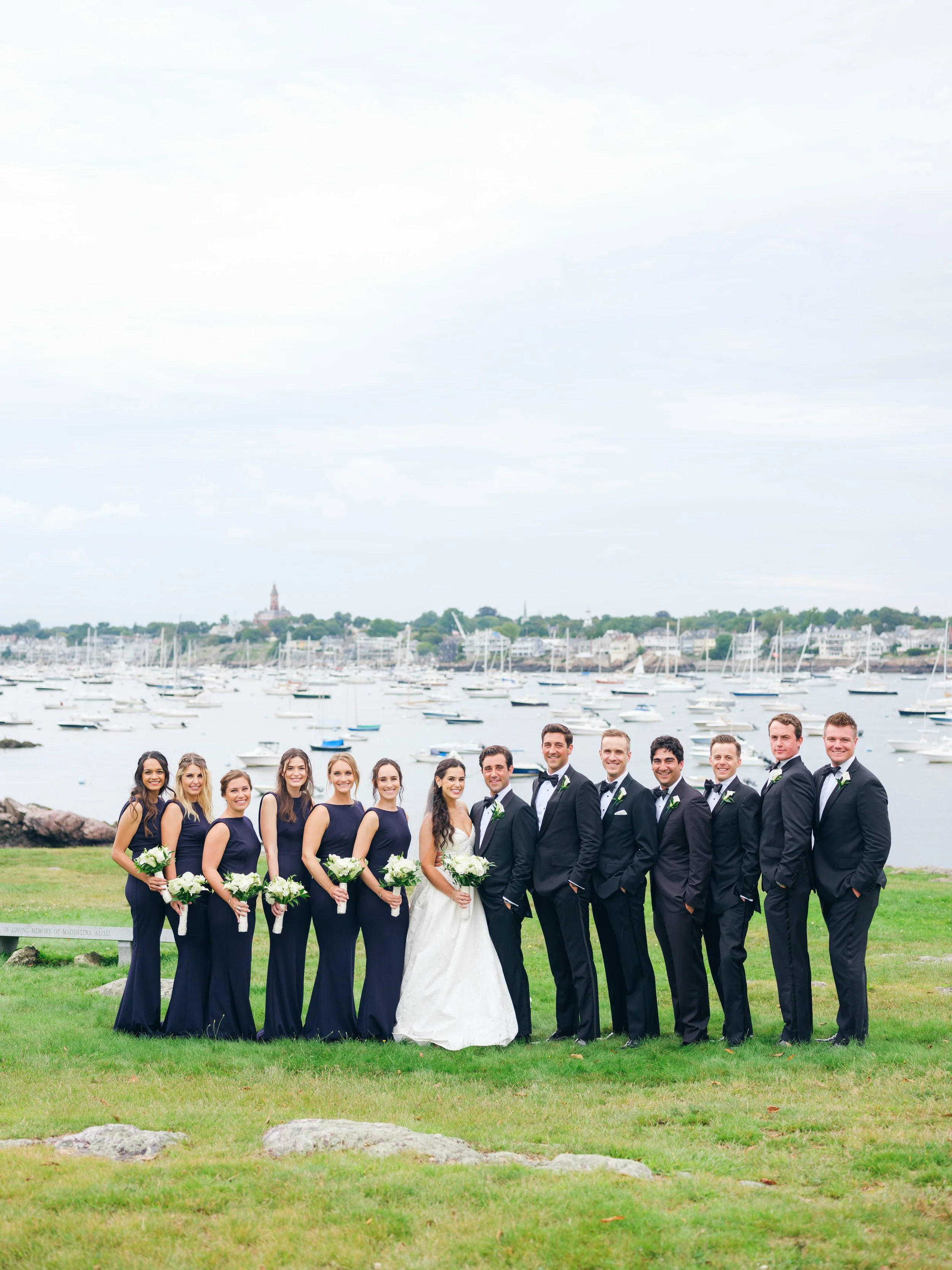 Group of 14 people dressed in formal attire, posing outdoors near a harbor with sailboats and a cityscape in the background.