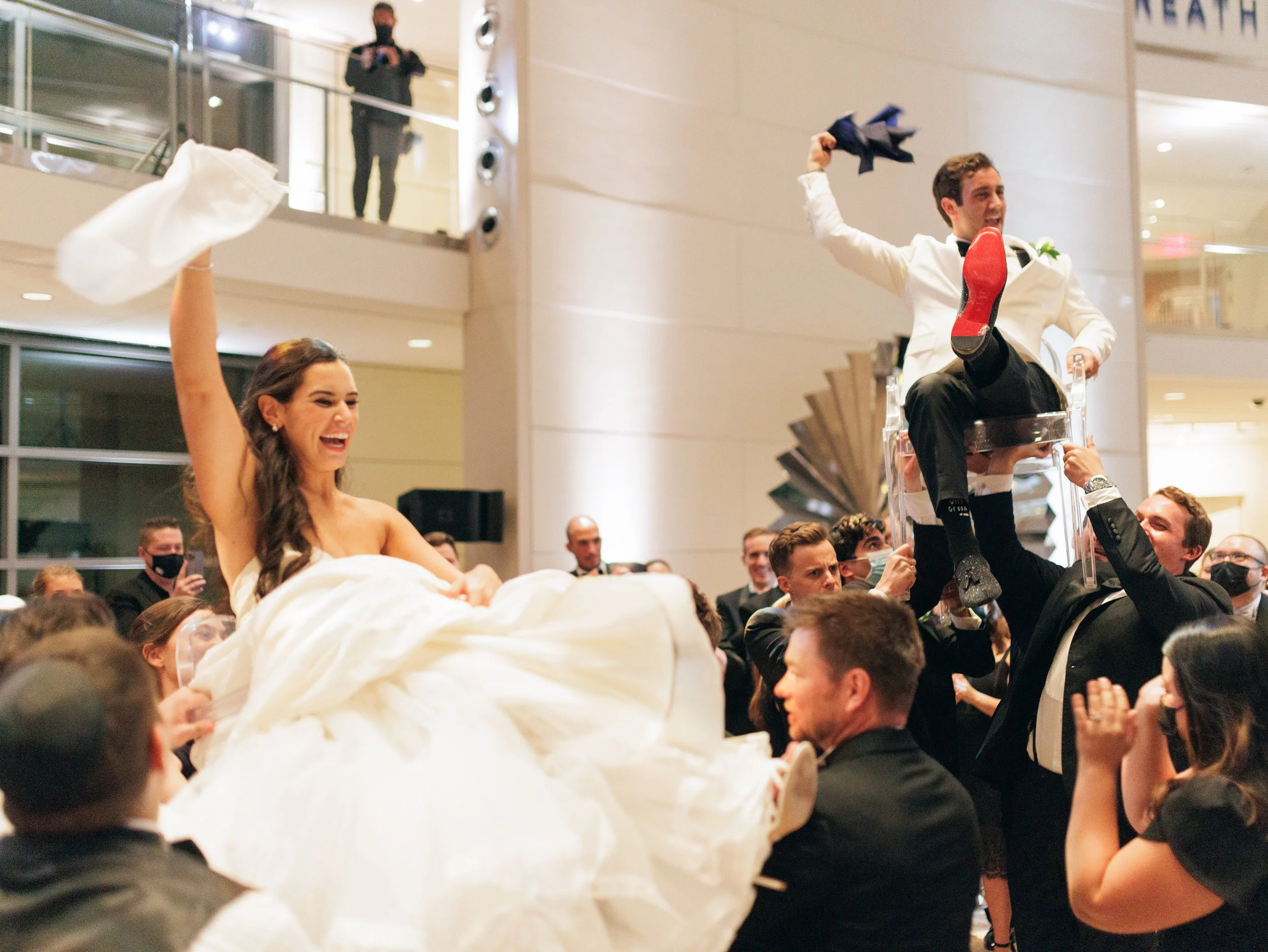 A couple dressed in formal attire, with the woman in a wedding dress and the man in a white tuxedo, riding on a large decorative float during a celebration or parade. The man around him is smiling and raising an arm, and a large crowd of onlookers su
