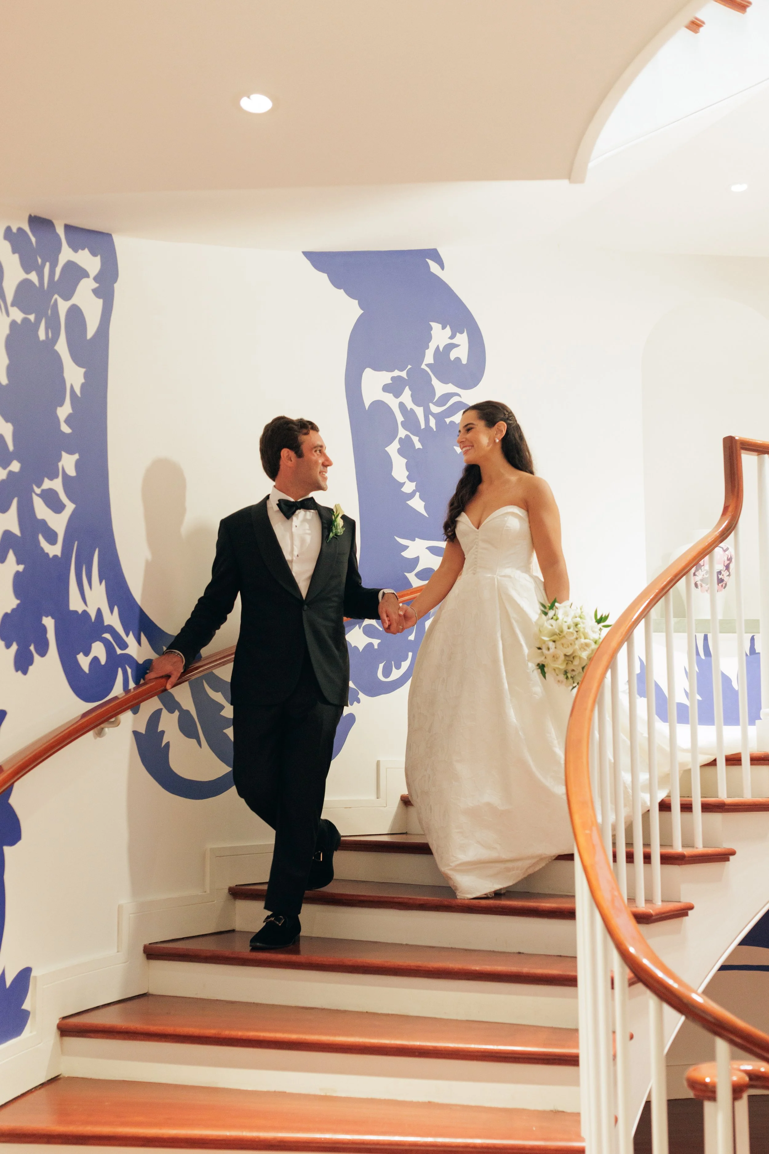 A smiling bride and groom holding hands on a staircase, with the groom in a tuxedo and the bride in a white wedding dress, near a blue floral mural.