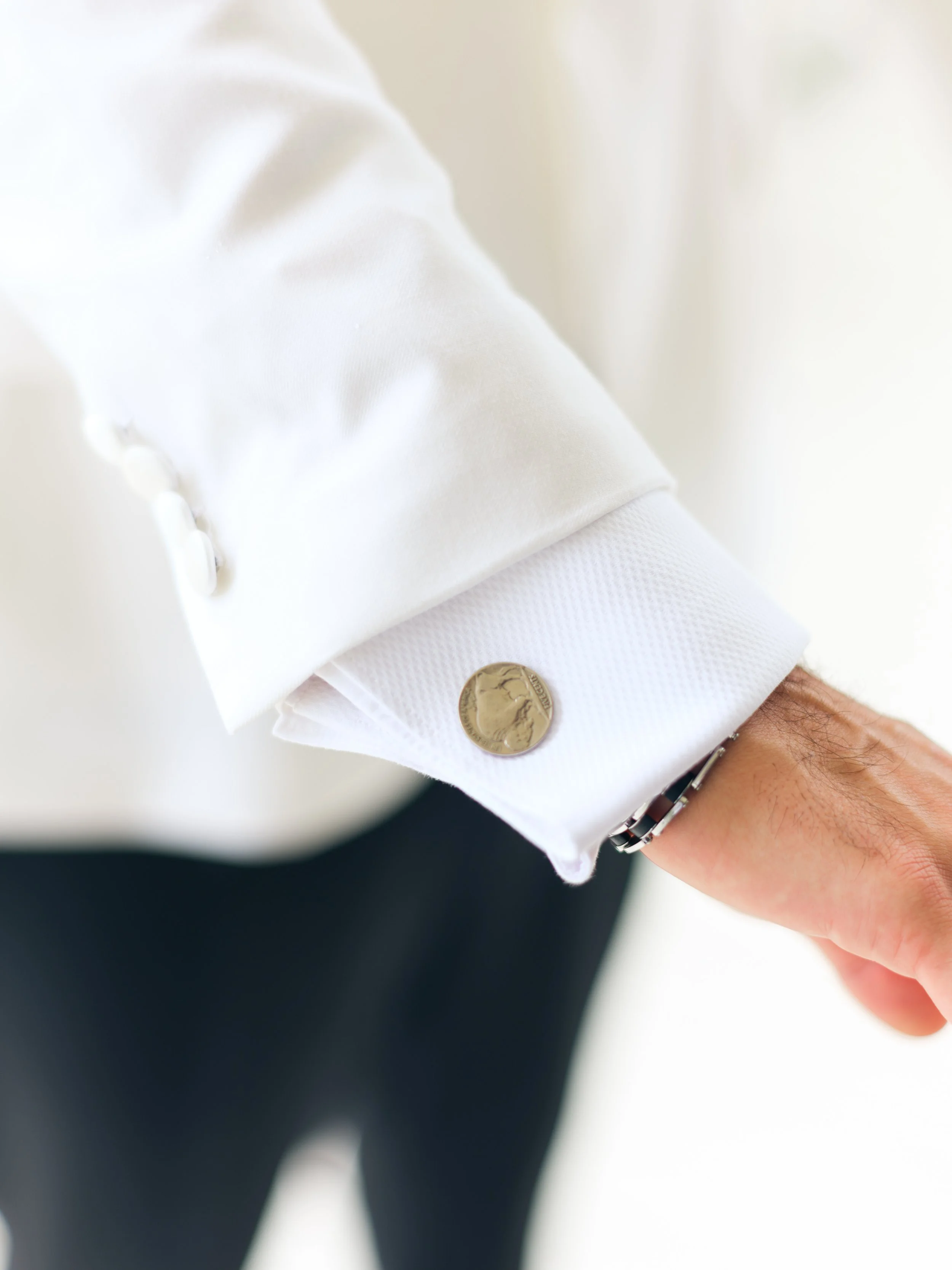 Close-up of a person's white shirt cuff with silver cufflinks, wearing a black bracelet, and showing a brown skin hand, blurred background.