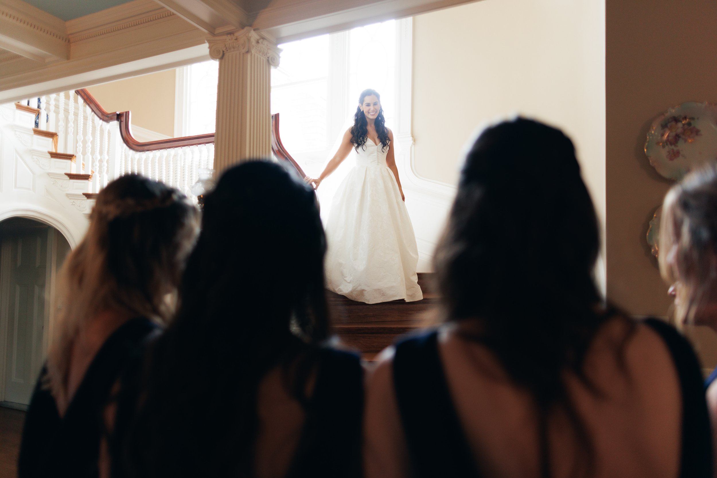 A bride in a white wedding dress standing at the top of a staircase, smiling, as guests watch her from below in a grand interior space with large windows and ornate pillars.