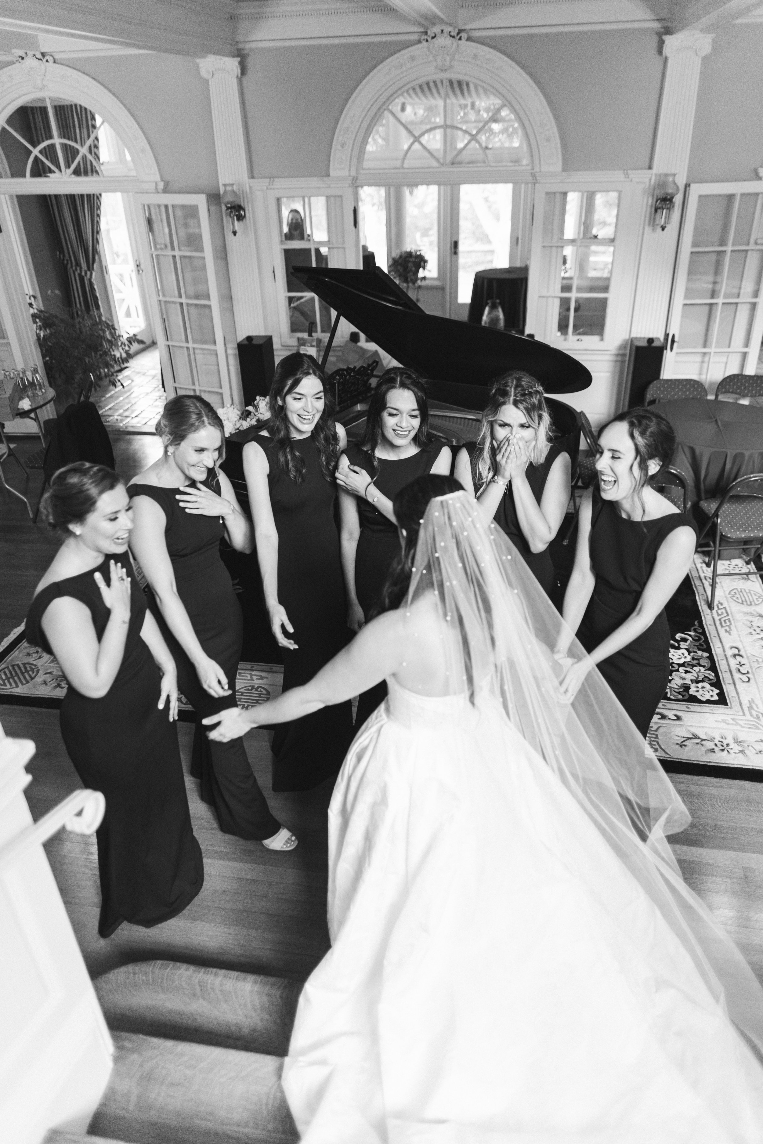 Group of six women in black dresses laughing and smiling during a wedding or special event, with a bride in a wedding gown and veil in the foreground.