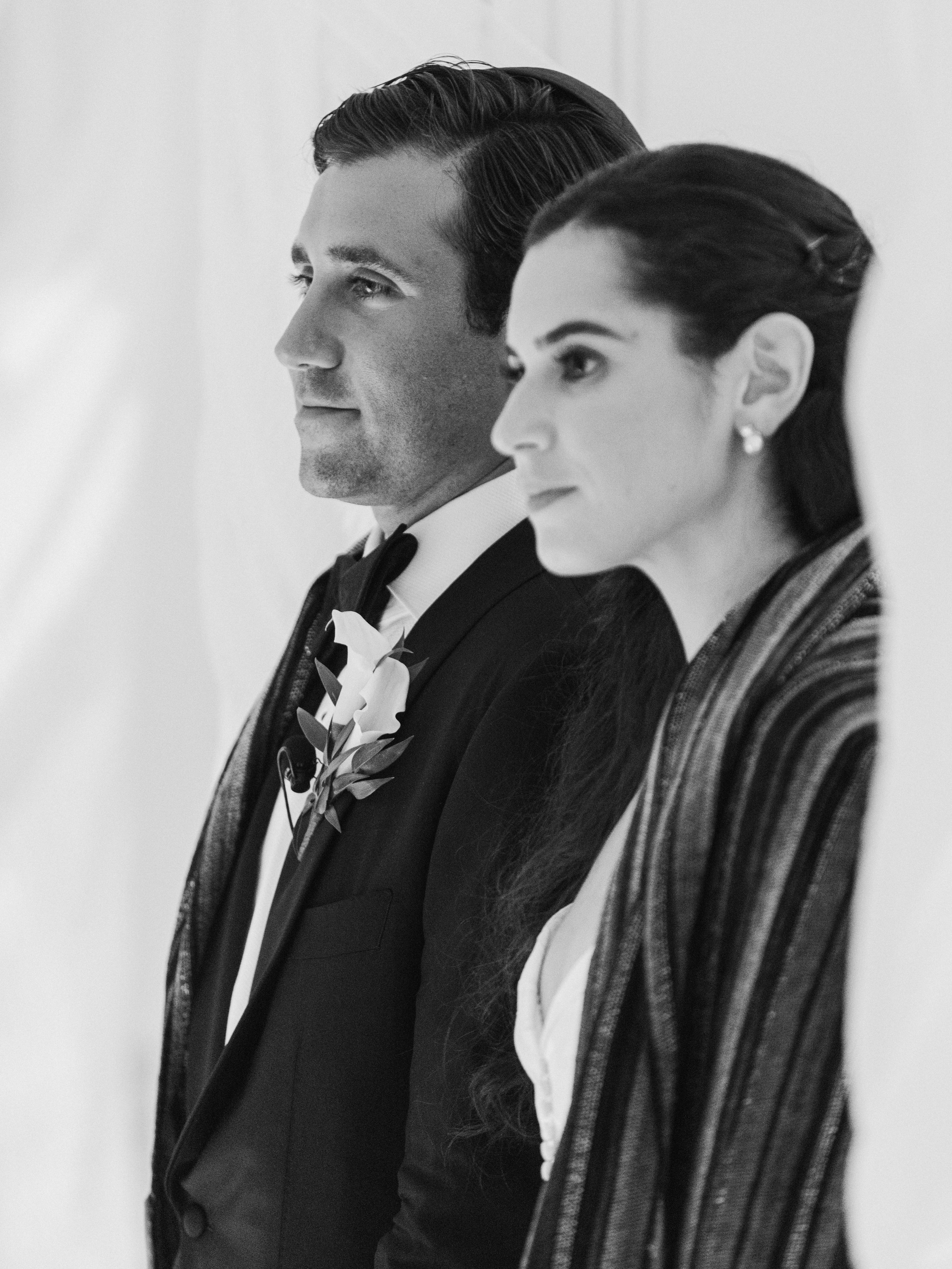 A black-and-white photo of a man and woman standing side by side, both with serious expressions, wearing formal attire, with the man in a tuxedo and the woman in a striped blazer.