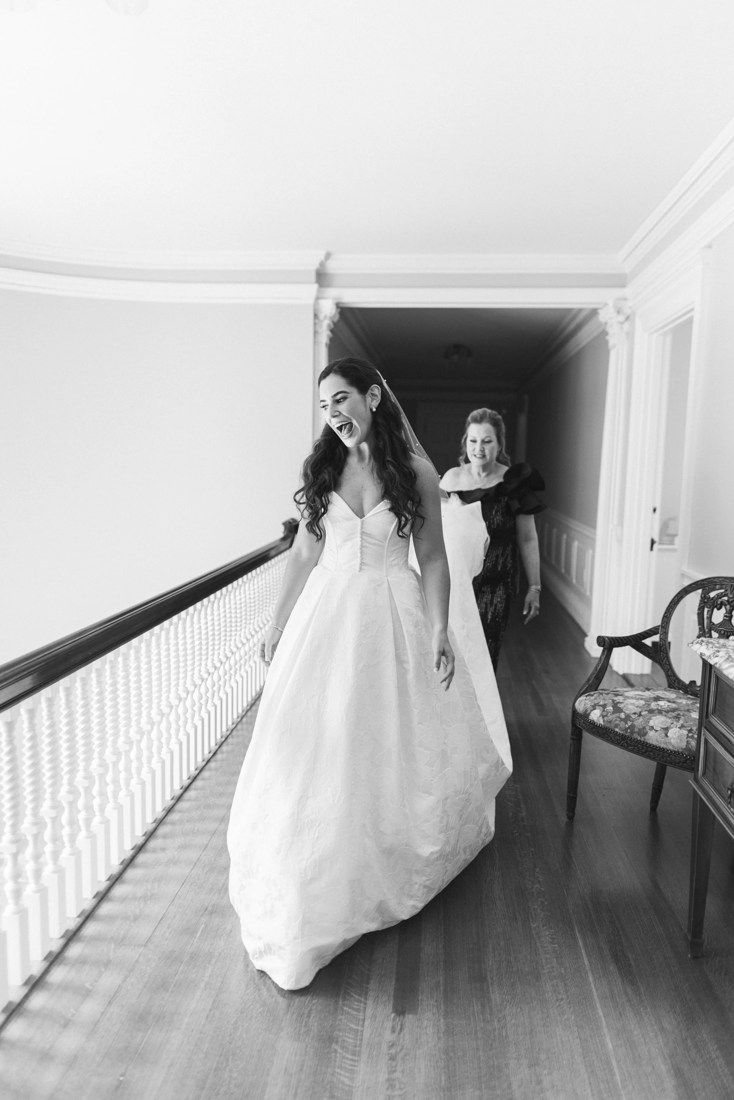 A bride in a strapless wedding gown with long dark hair walking in a hallway, smiling, with a woman in a dark dress following her. The hallway has ornate crown molding and a wooden floor, with chairs along the wall.