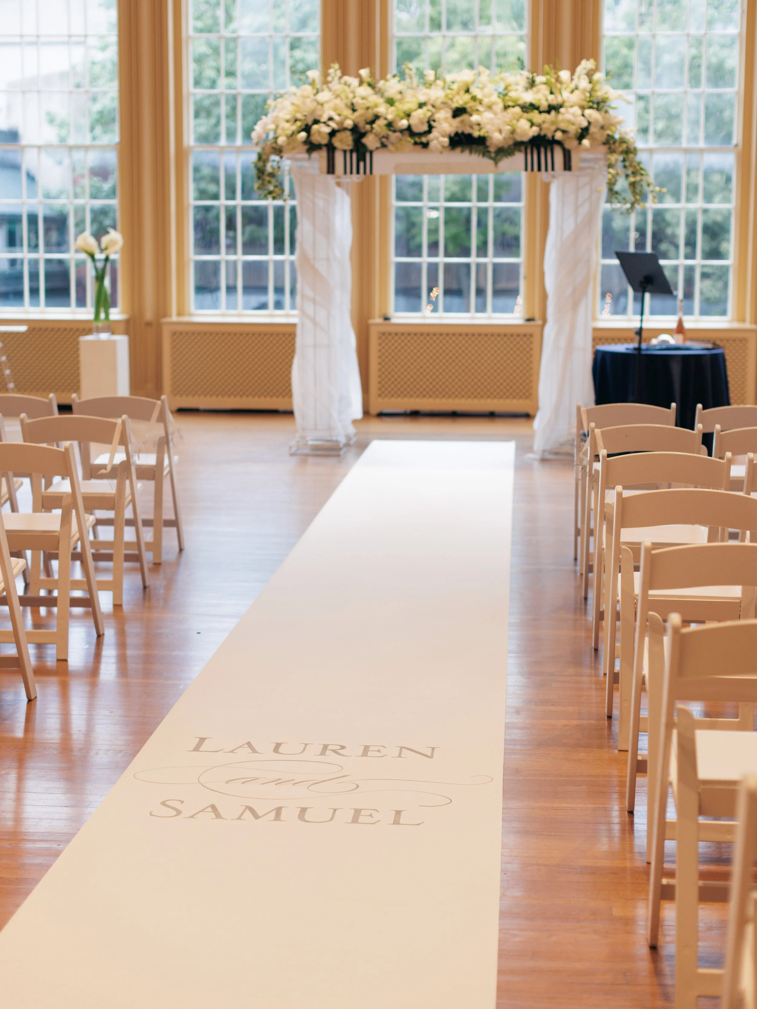 Wedding ceremony setup with a white aisle runner that has the names Lauren and Samuel printed on it. The aisle is flanked by white chairs, leading to an arch decorated with white flowers, situated in front of large windows in a bright room.
