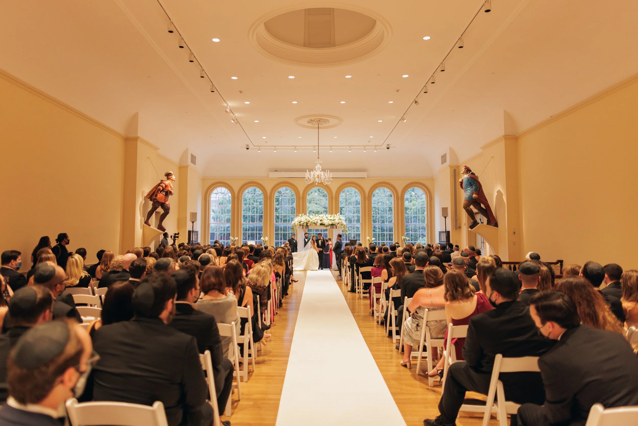 A large wedding ceremony inside a spacious, elegant hall with high arched windows and a chandelier. The bride and groom are standing under a floral arch, surrounded by friends and family seated on white chairs. Two large statues of clowns are positio