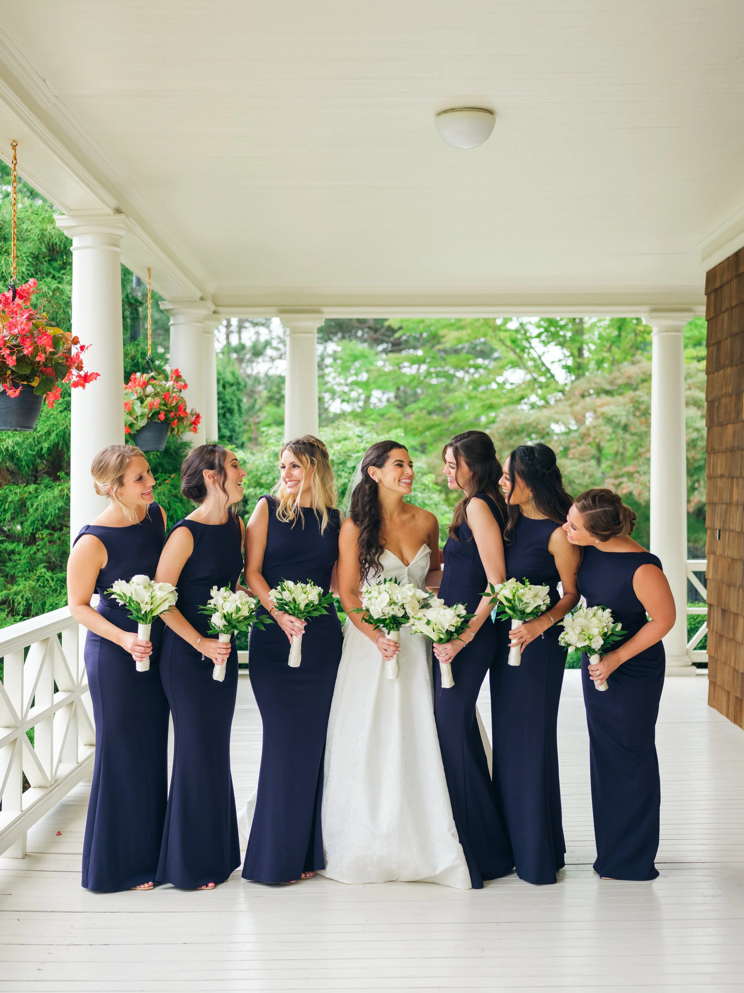 A bride and six bridesmaids on a porch with white columns and lush green trees in the background. The bridesmaids are wearing long navy blue dresses and holding white bouquets, and the bride is in a white wedding gown holding a larger white bouquet. 