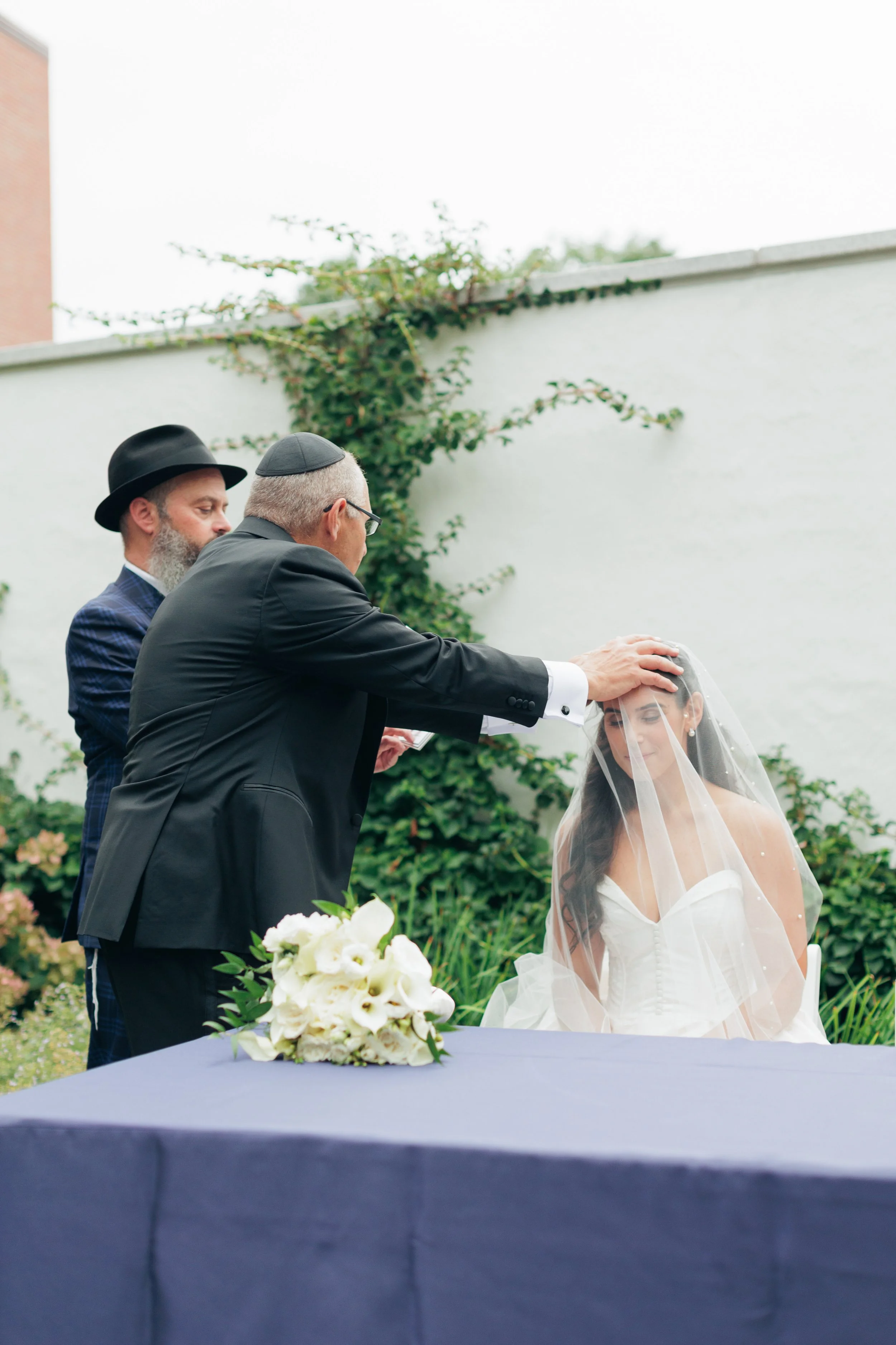 A wedding ceremony outdoors with a bride wearing a white dress and veil, and a groom in a black suit. Two men, one with a beard and hat, and the other with gray hair and kippah, are placing a hand on the bride's head. A bouquet of white flowers is on