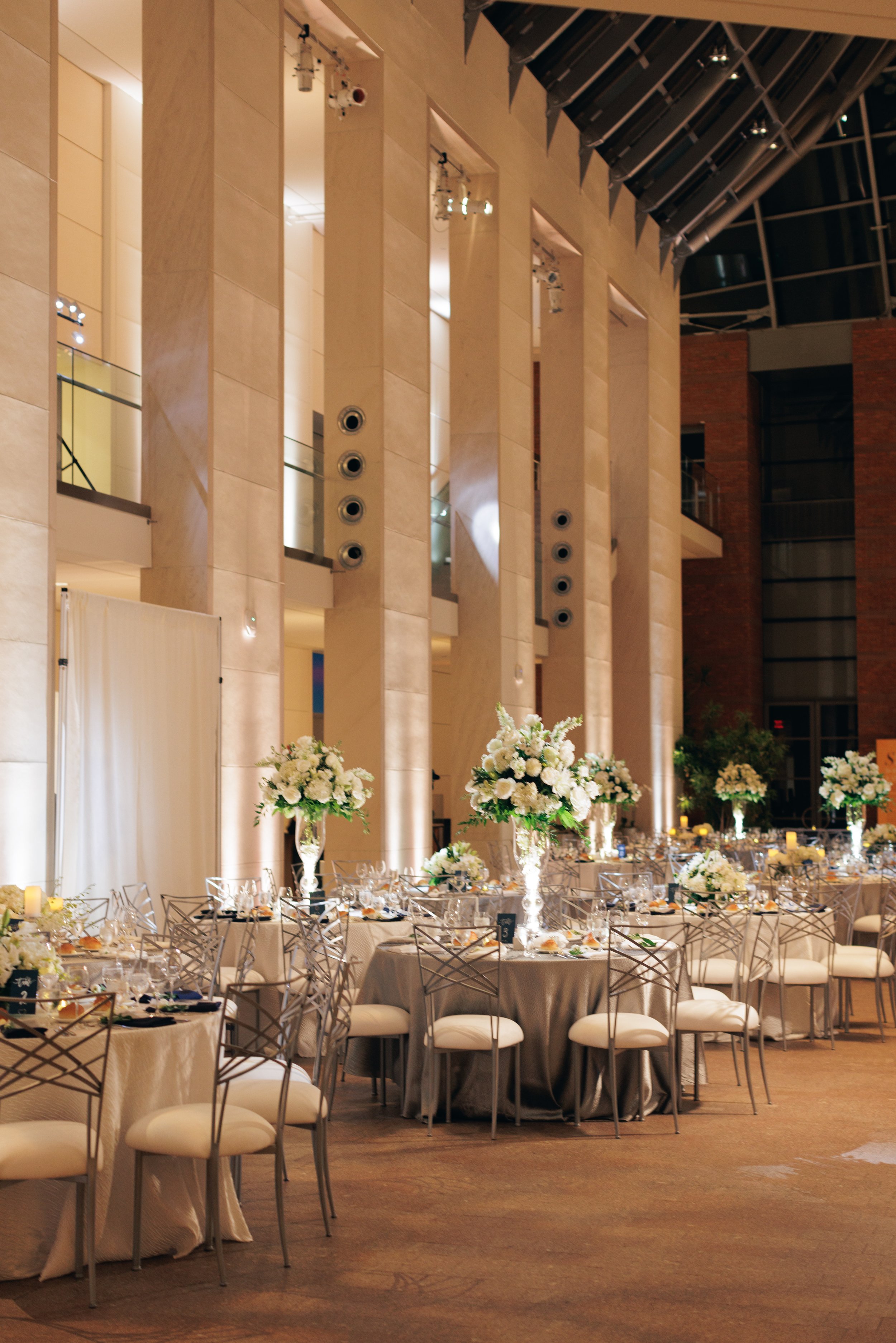 Elegant banquet hall with round tables decorated with white floral centerpieces, set with glassware and dinnerware, surrounded by white chairs with metal frames, with large columns and high ceiling featuring exposed metal beams.