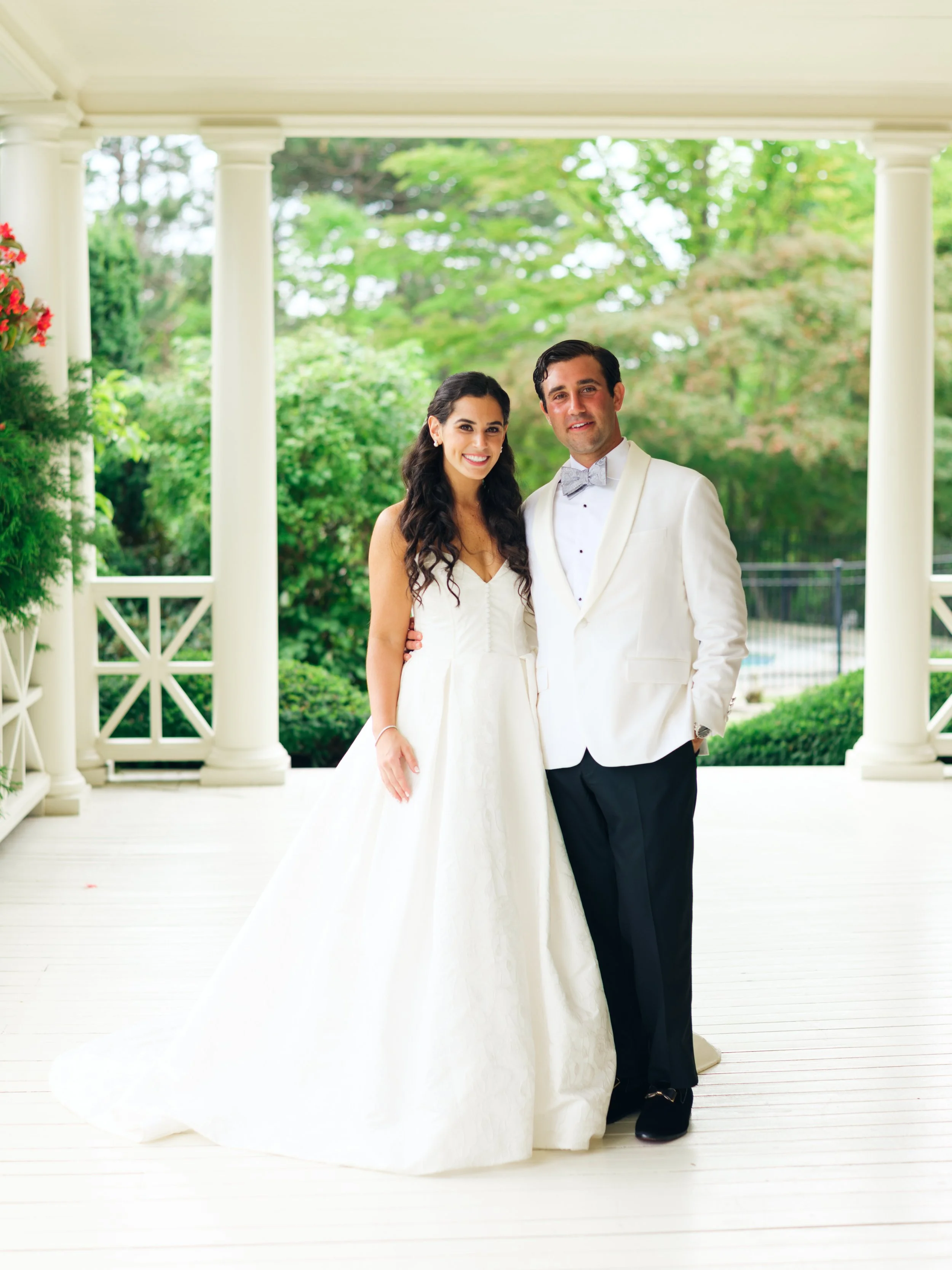 A newlywed couple stands together on a white porch with white columns and green trees in the background. The woman wears a white wedding dress, and the man wears a white tuxedo with a bow tie.