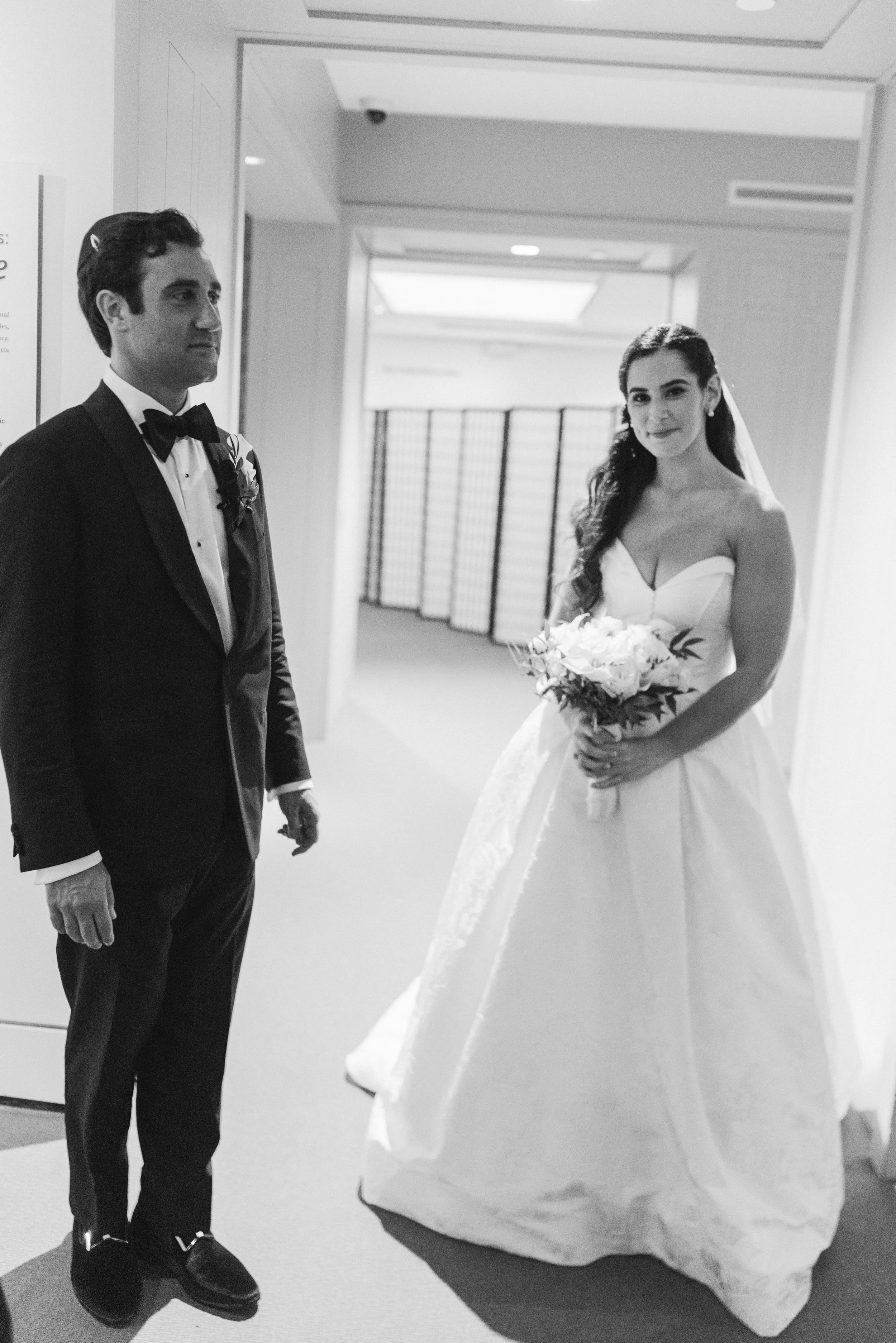 A man in a tuxedo and a woman in a wedding dress standing in an indoor space, possibly a hallway or lobby. The woman is holding a bouquet of flowers and smiling at the camera.