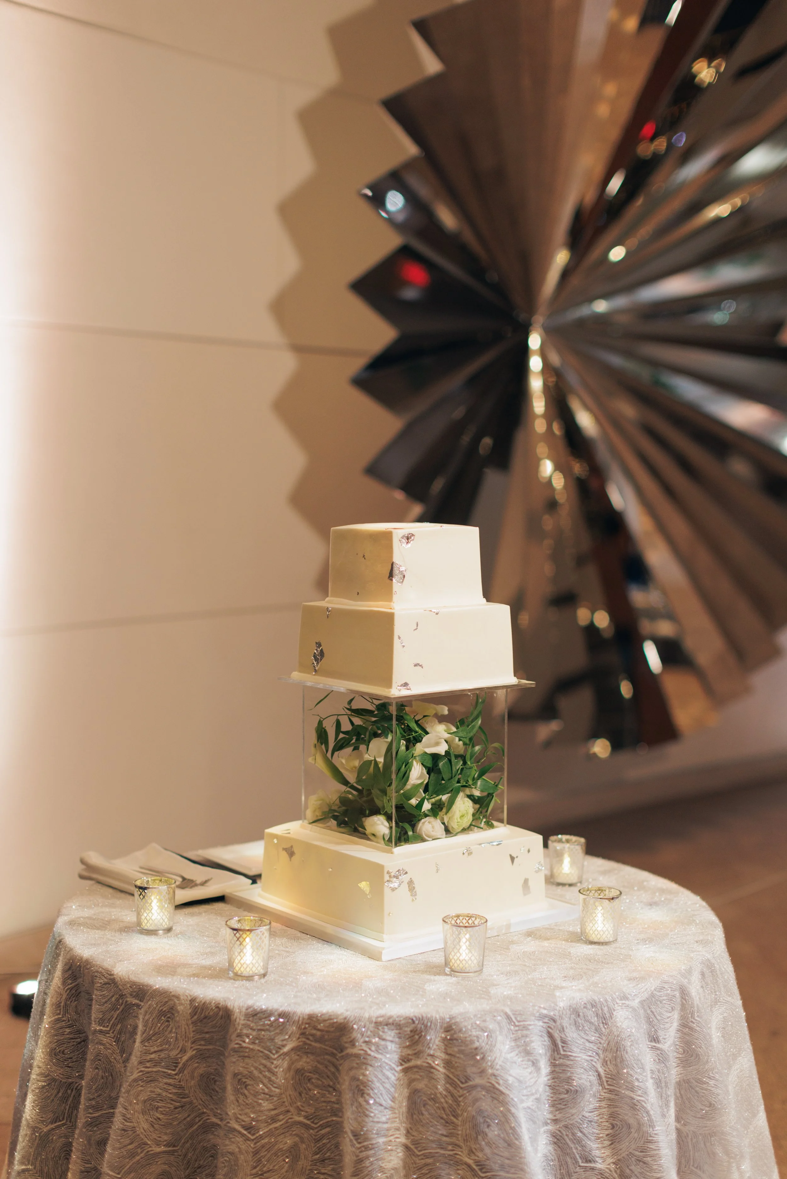 A tiered wedding cake with white fondant, silver leaf accents, and fresh white flowers, displayed on a table with a lace tablecloth and surrounded by votive candles. In the background, a large decorative wooden installation with geometric shapes is v