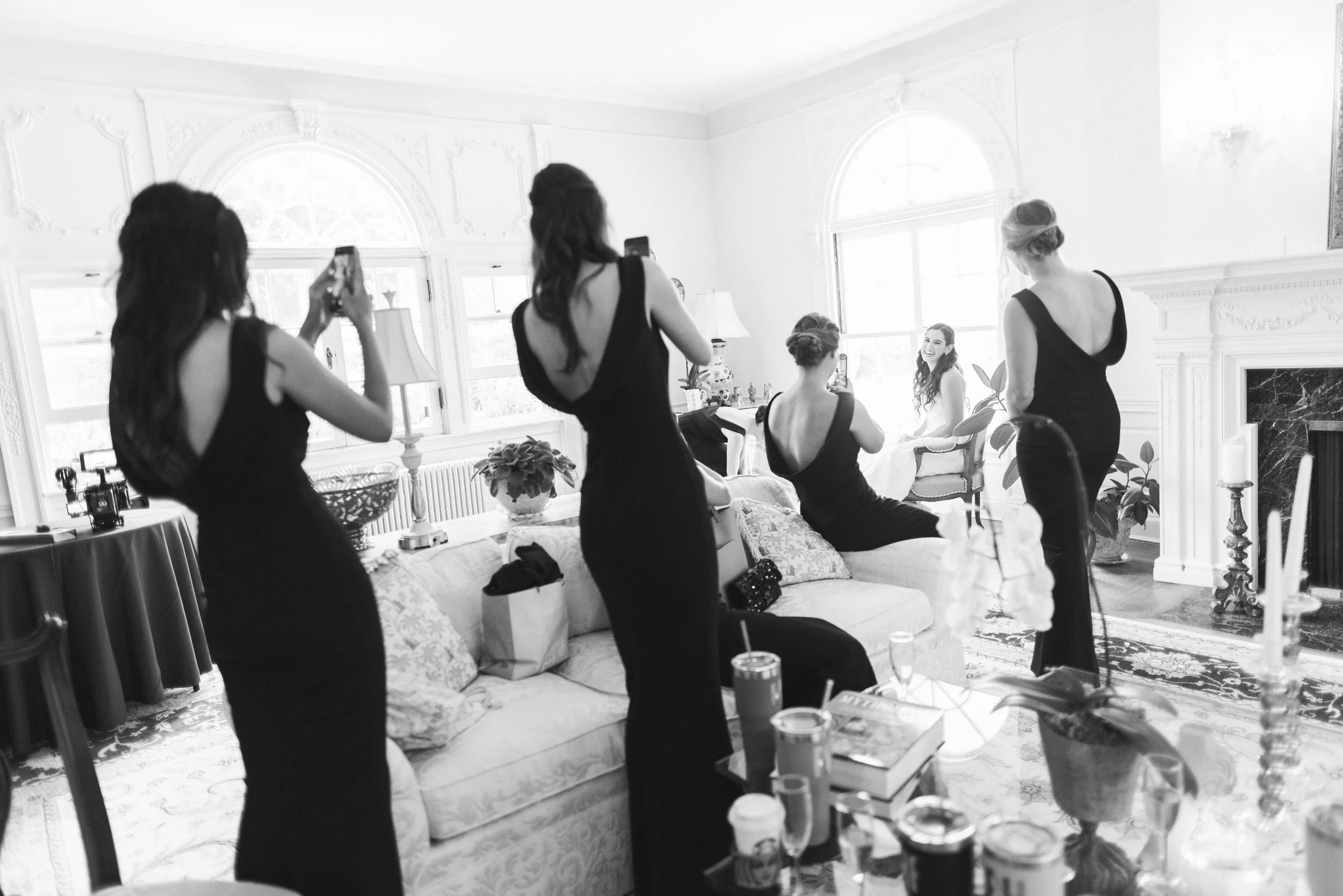 Women in black dresses getting ready and taking photos in a bright, elegant living room with large windows, ornate decorations, and a classical style fireplace.