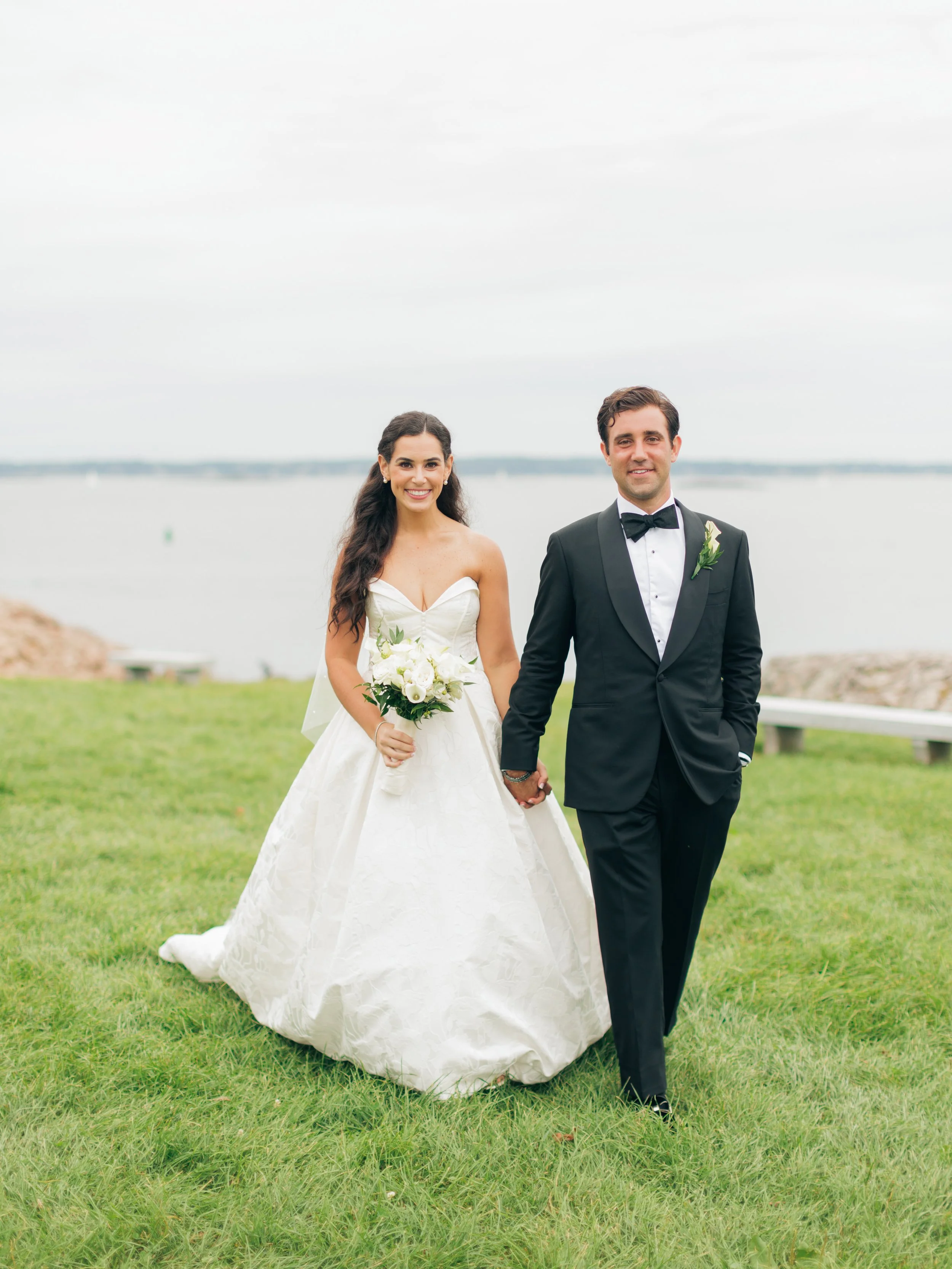 A newlywed couple standing on a grassy field near a body of water, dressed in wedding attire, holding hands and smiling.