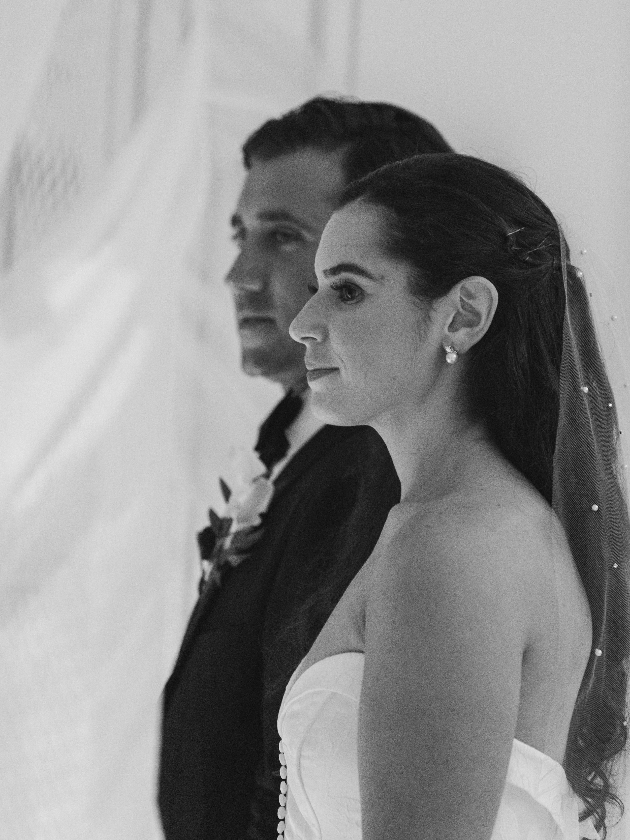 A black-and-white photo of a bride and groom standing close, with their sides to the camera, sharing a quiet moment.