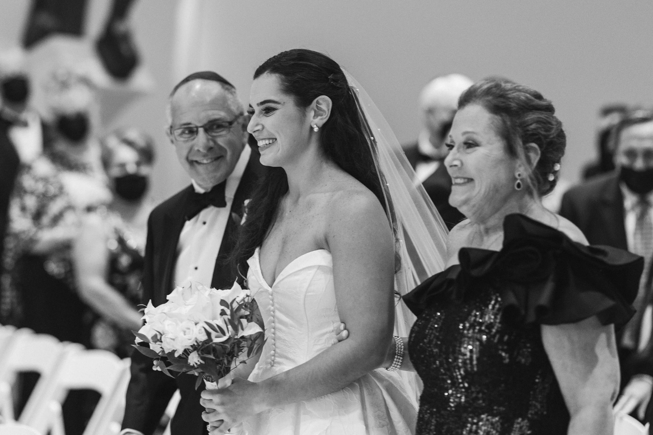 A bride in a white wedding dress holding a bouquet, smiling as she exchanges vows at her wedding ceremony. She is surrounded by her parents and family members, who are also smiling.