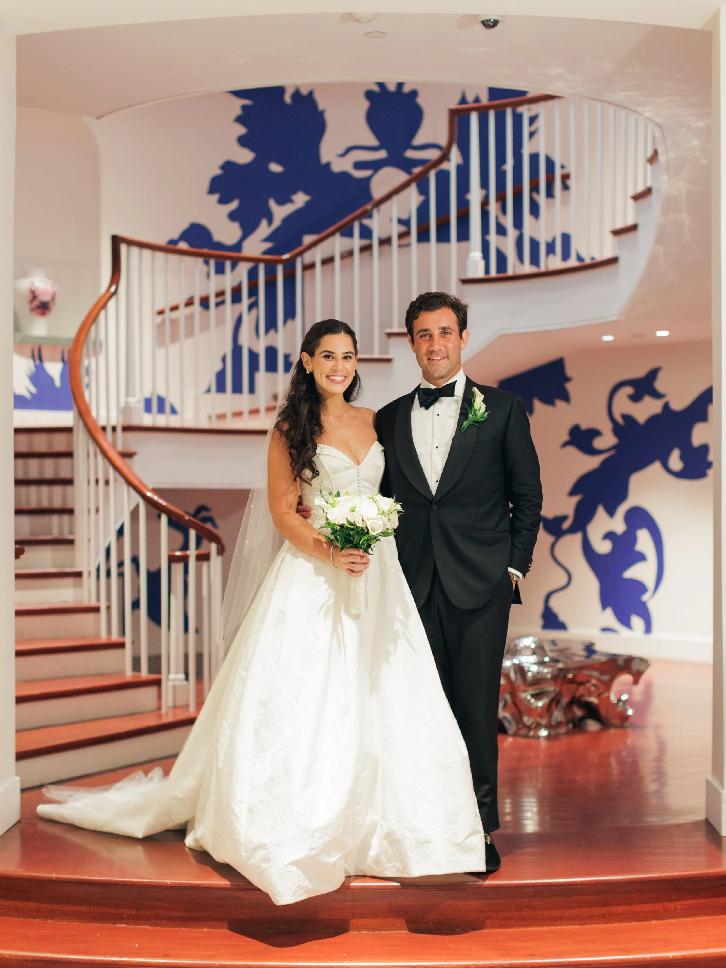 Bride and groom in wedding attire standing on a curved staircase with blue decorative wall art behind them.