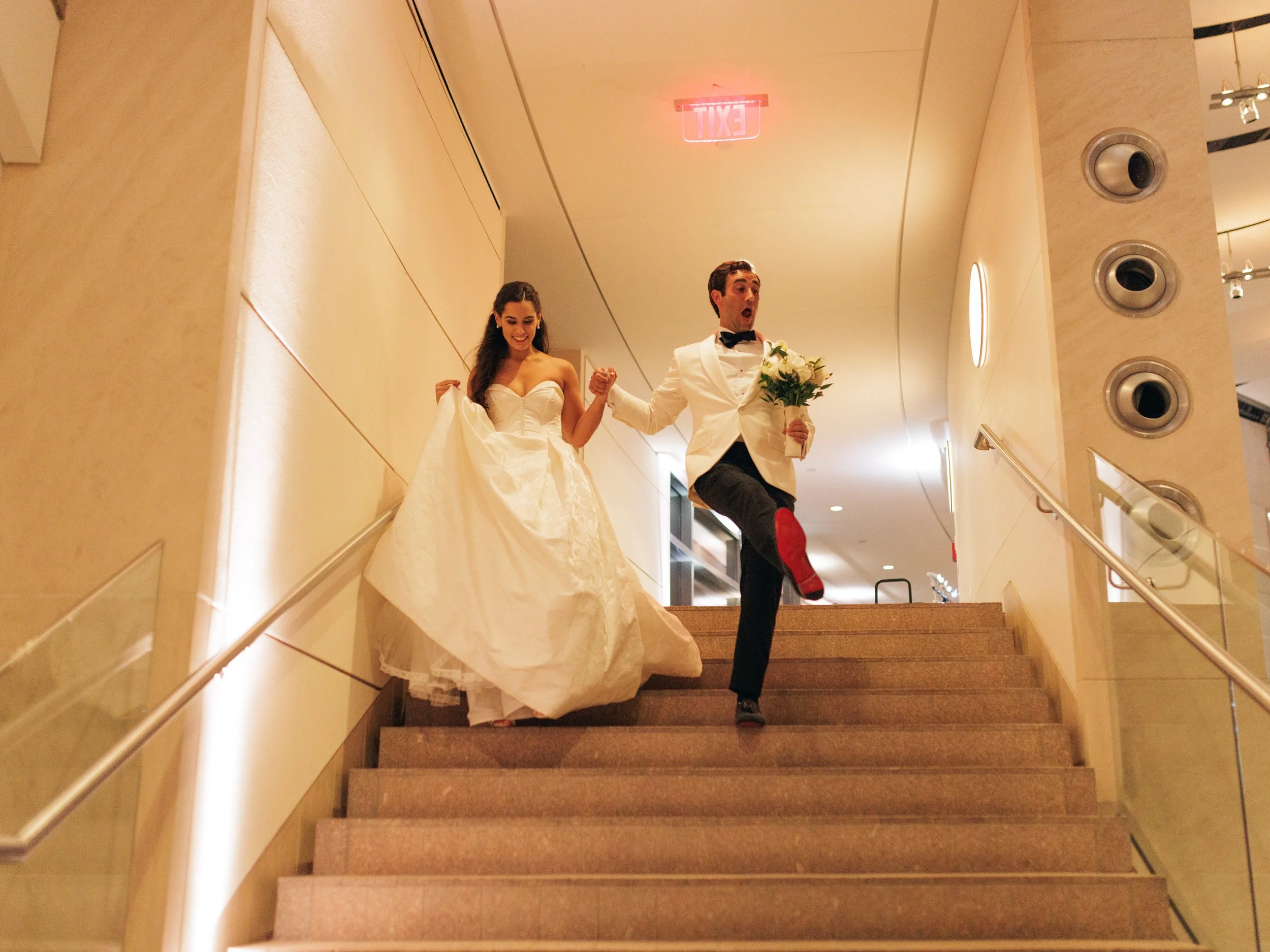 A bride and groom walking down a staircase, holding hands. The bride is wearing a white wedding dress and the groom is wearing a white tuxedo jacket with black pants, a black bow tie, and red shoes. The bride is smiling and holding part of her dress,