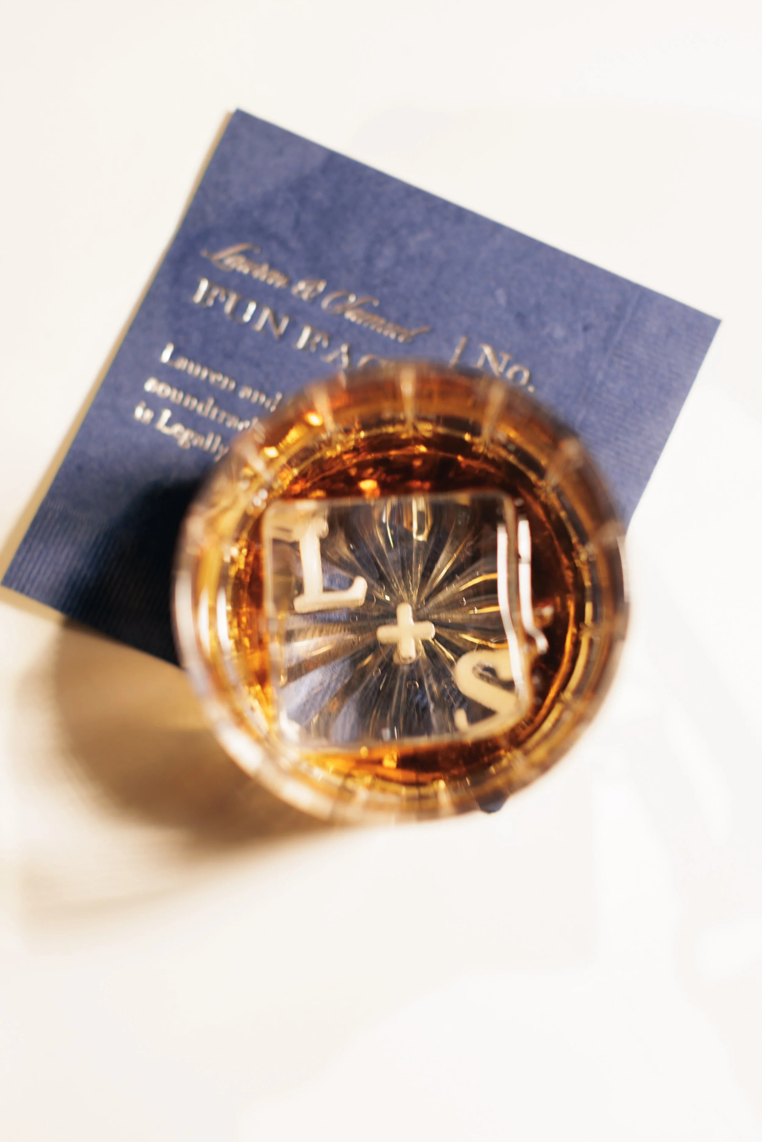 Close-up view looking down into a glass of amber-colored liquid on a white surface with a blue paper underneath, showing embossed silver text.