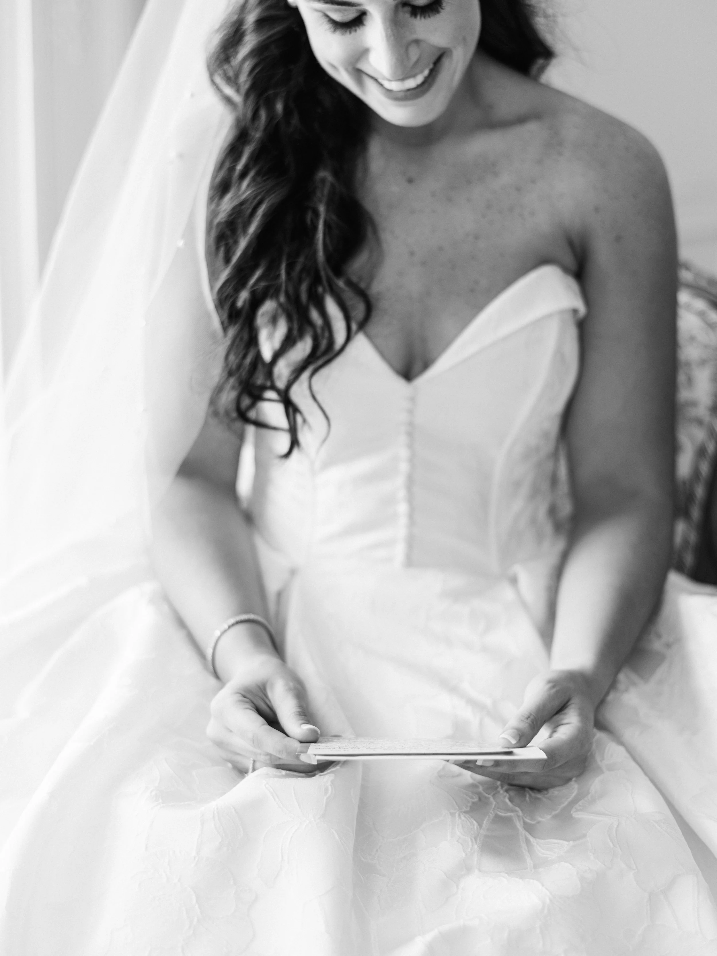 Black and white photo of a woman sitting on a bed, smiling, looking down at a phone she is holding with both hands. She has long, wavy hair and is wearing a strapless dress.