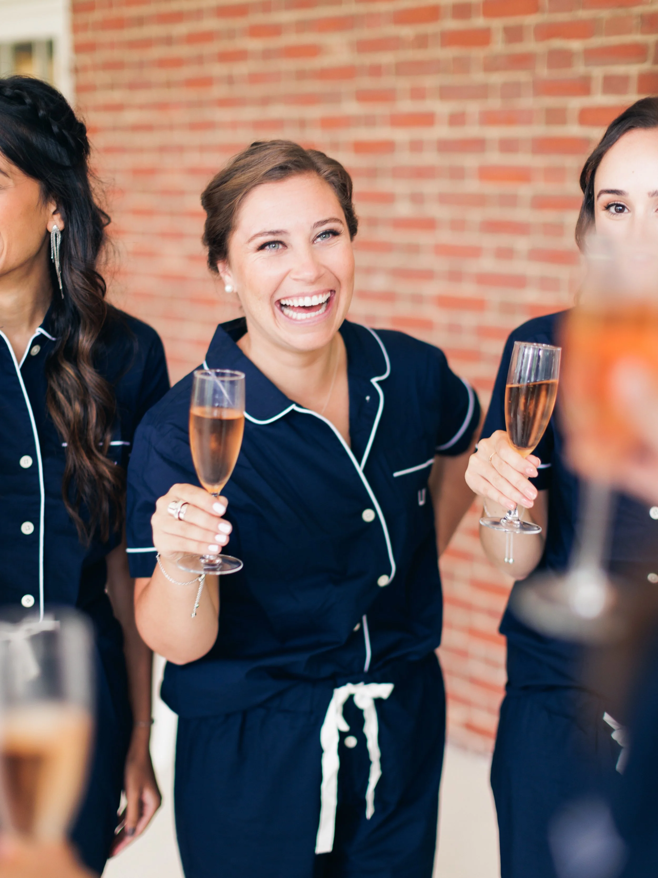 A woman in navy pajamas with white piping holding a glass of champagne, smiling and laughing, at a social gathering.