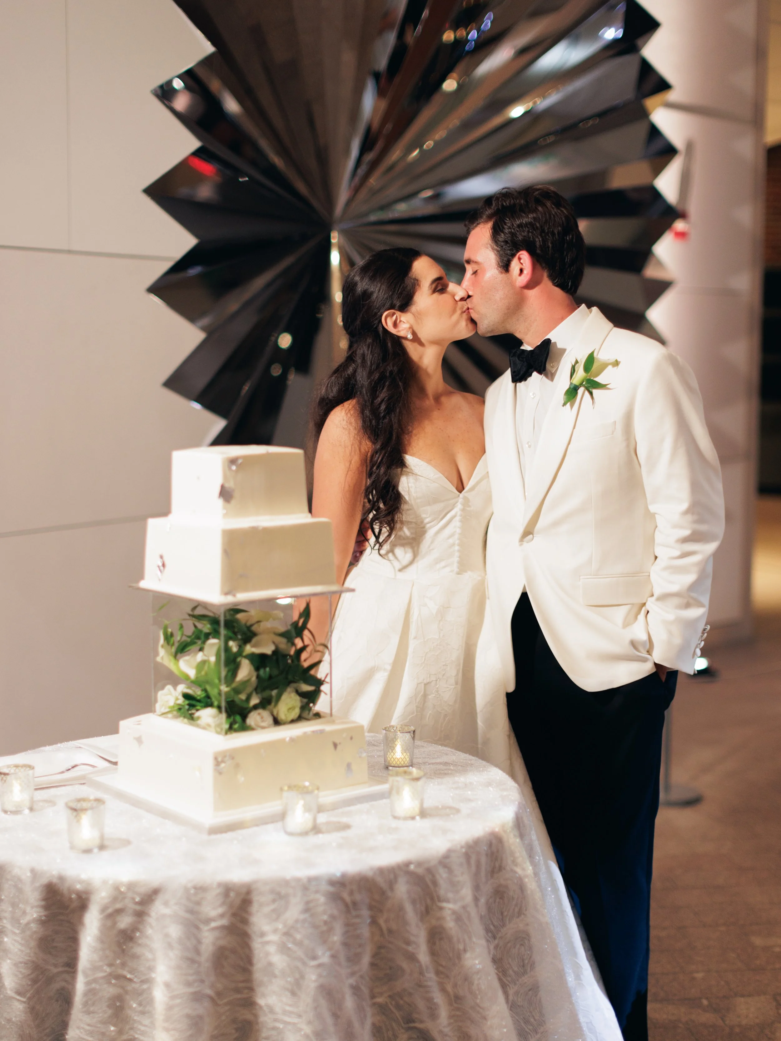 A bride and groom kissing at their wedding reception, with a wedding cake and floral decorations on the table in front of them, and a large decorative mirror behind them.