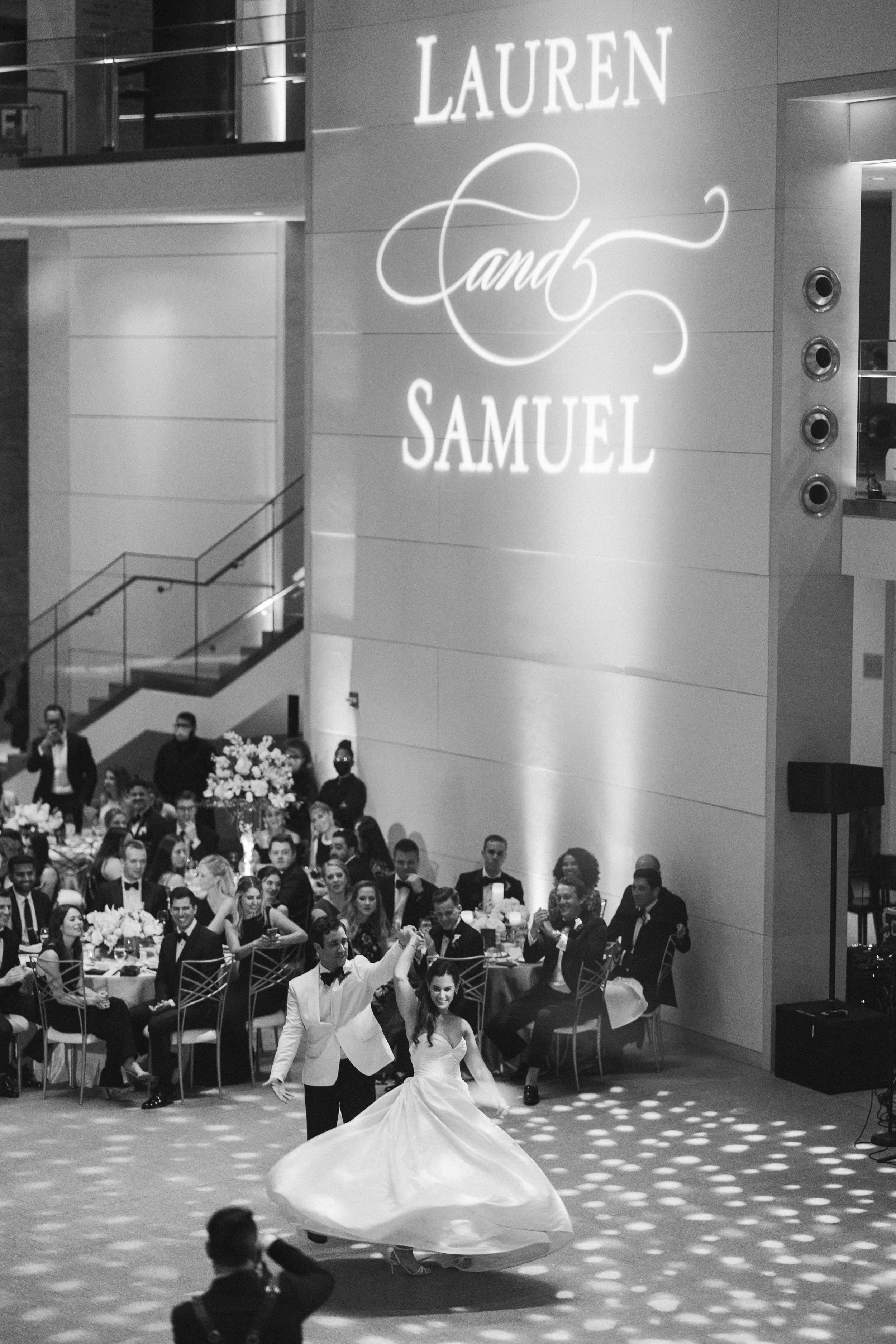 A couple in wedding attire dancing in front of a large seated audience at a formal event. The room is decorated elegantly, with floral arrangements and tables set for guests. There is a large illuminated sign on the wall that says "LAUREN Sand SAMUEL