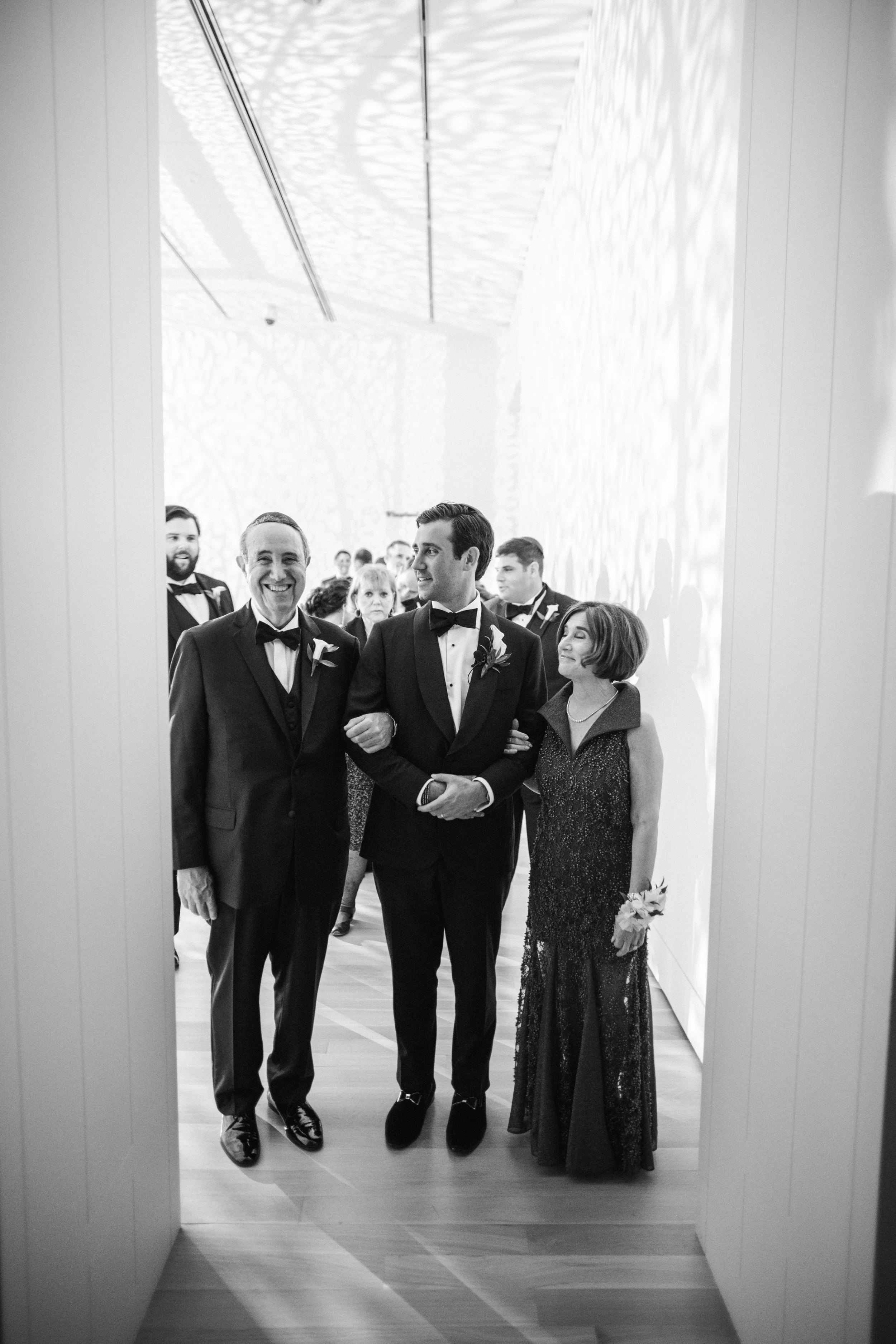Black and white photo of a wedding procession with a man in a tuxedo and a woman in a gown, walking down a hallway with onlookers watching.