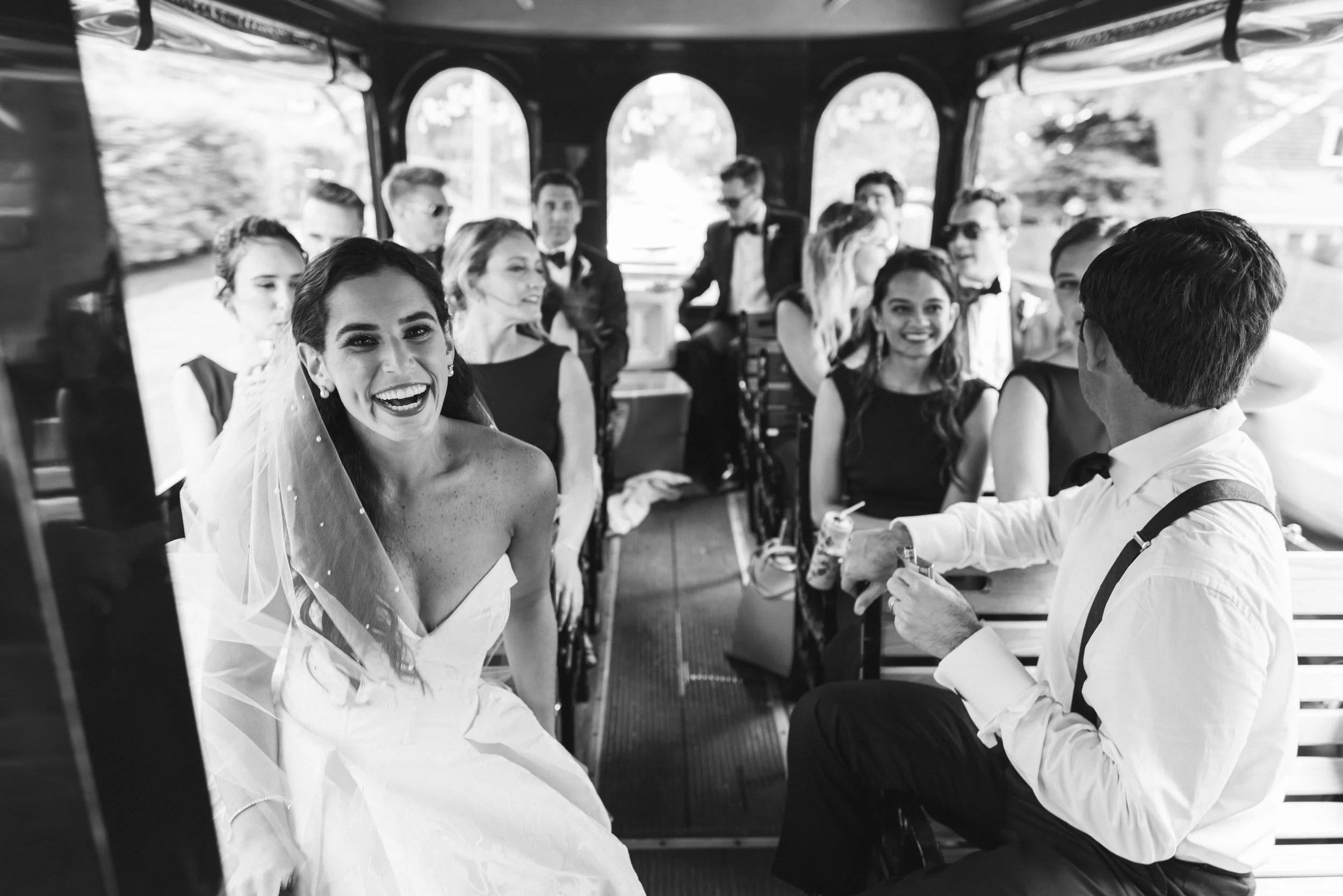 A group of people dressed in formal attire, including a woman in a wedding dress, on a trolley or bus with large windows and trees outside.