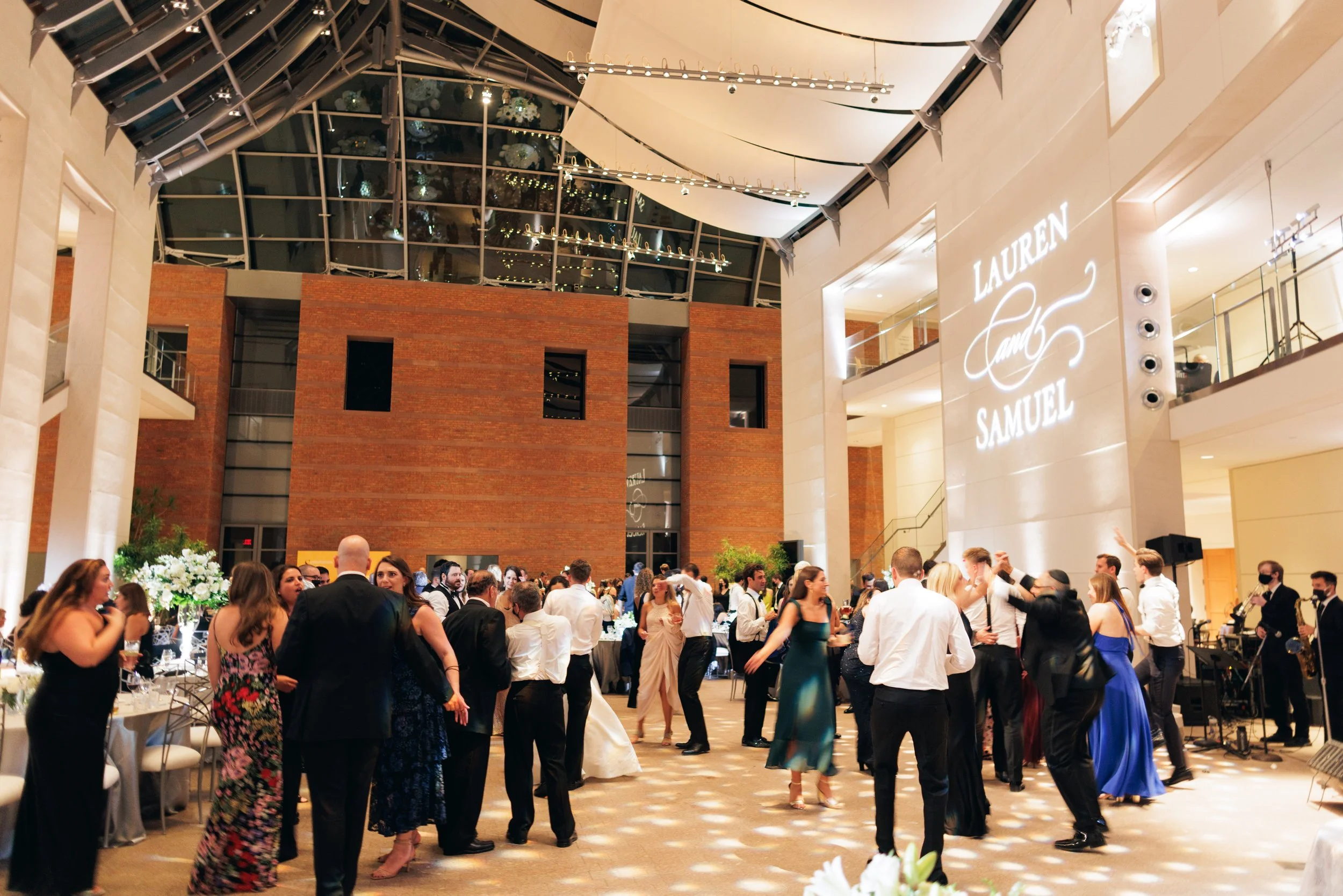 A grand indoor event with guests dancing and socializing, decorated with floral centerpieces, at the Lauren and Samuel wedding celebration.