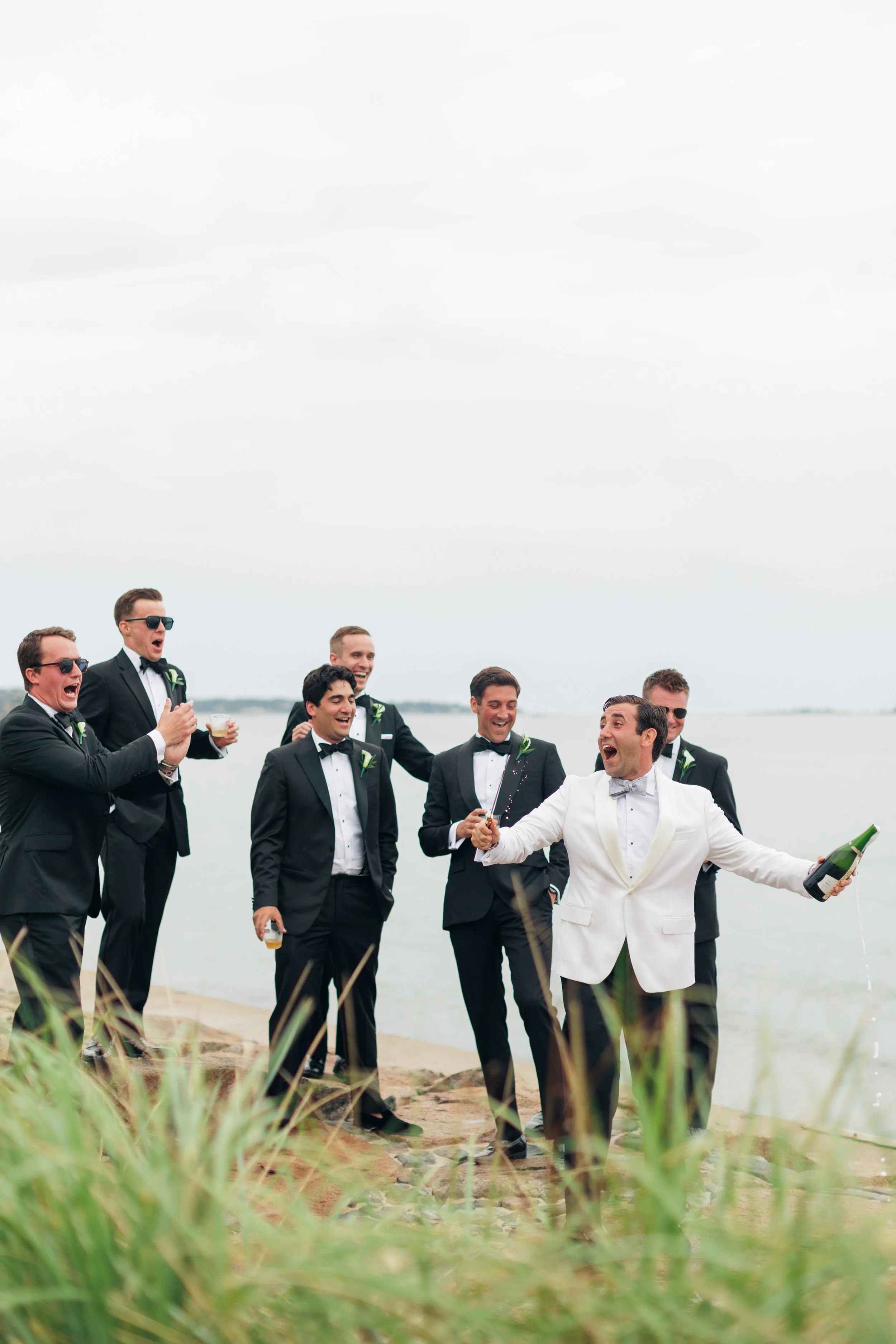 A group of men dressed in tuxedos celebrating on a beach, with some holding drinks and champagne. The scene appears joyful and festive, with a cloudy sky and water in the background.