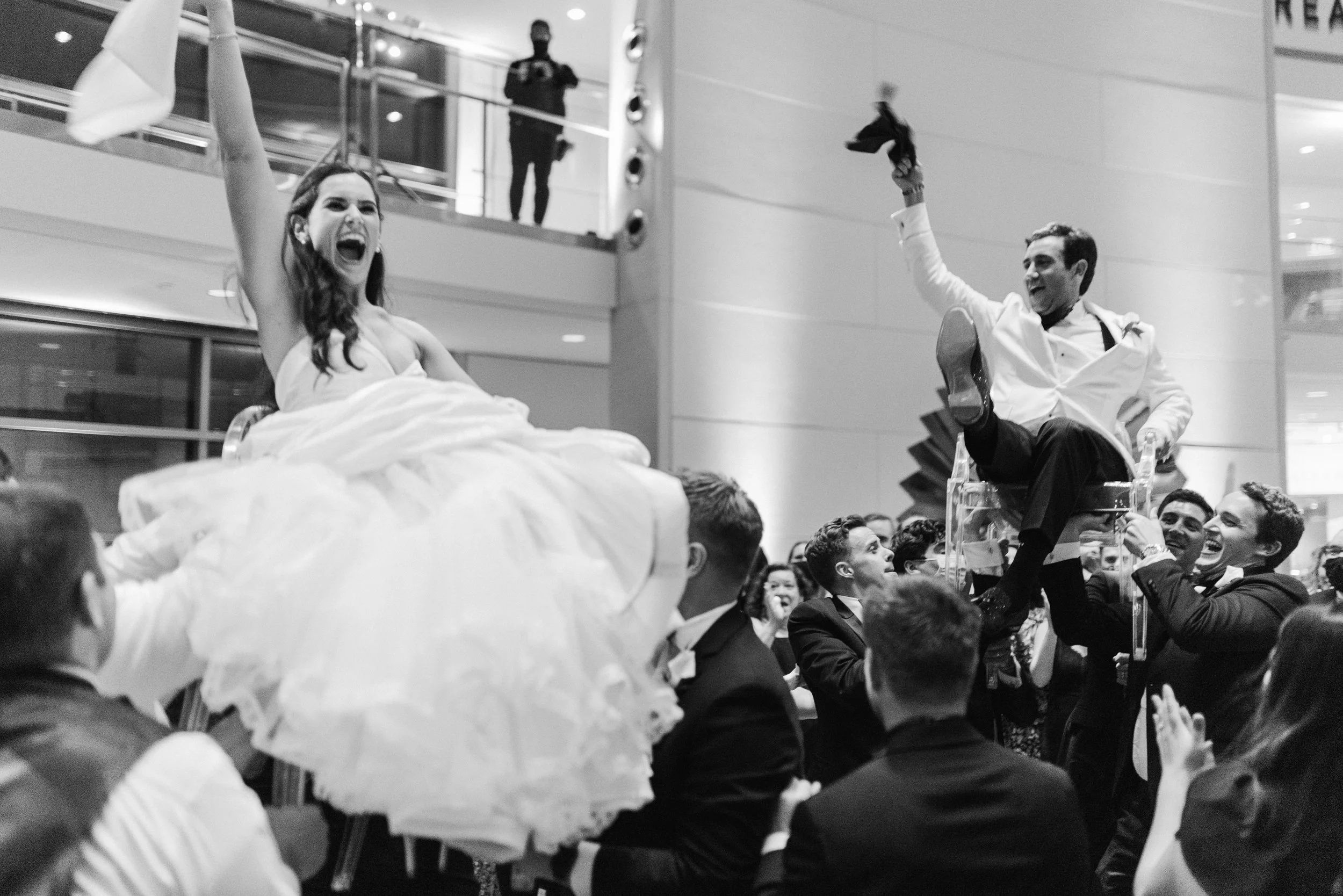 A woman in a wedding dress and a man in formal attire celebrating on a cake at a wedding reception, surrounded by guests. The scene is lively and joyous, with people smiling and cheering.