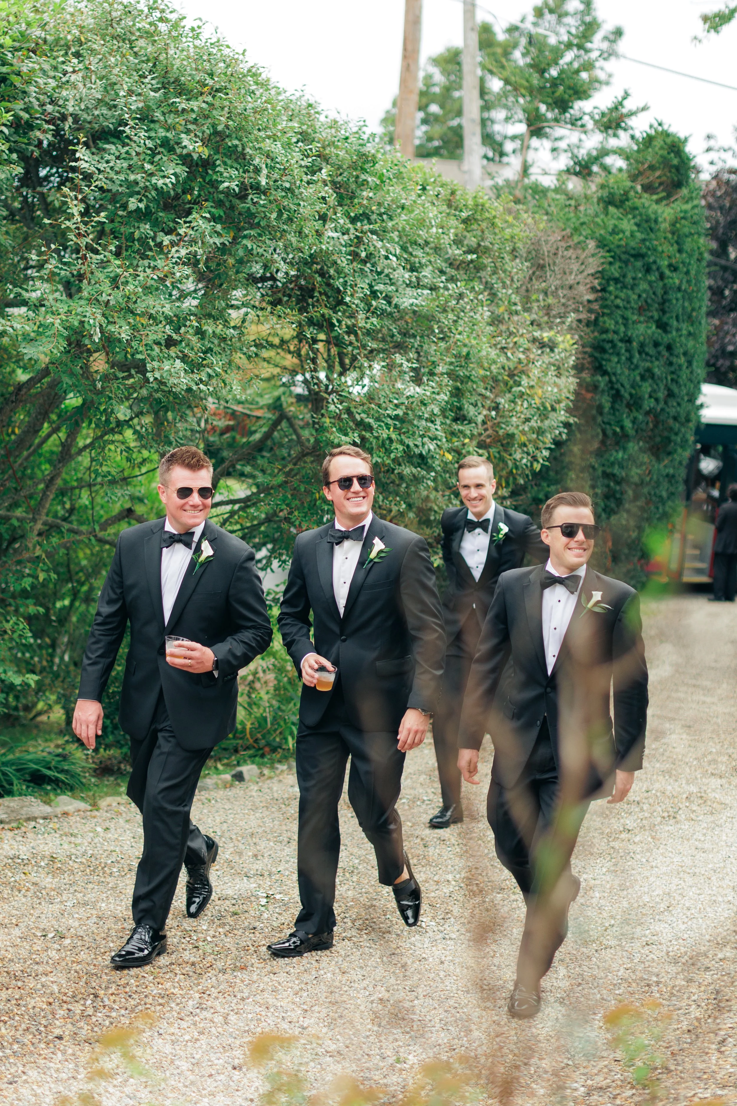 Four men dressed in black tuxedos with white shirts and black bow ties, wearing sunglasses and boutonnières, walking outdoors on a gravel path surrounded by trees and greenery, with some holding drinks.