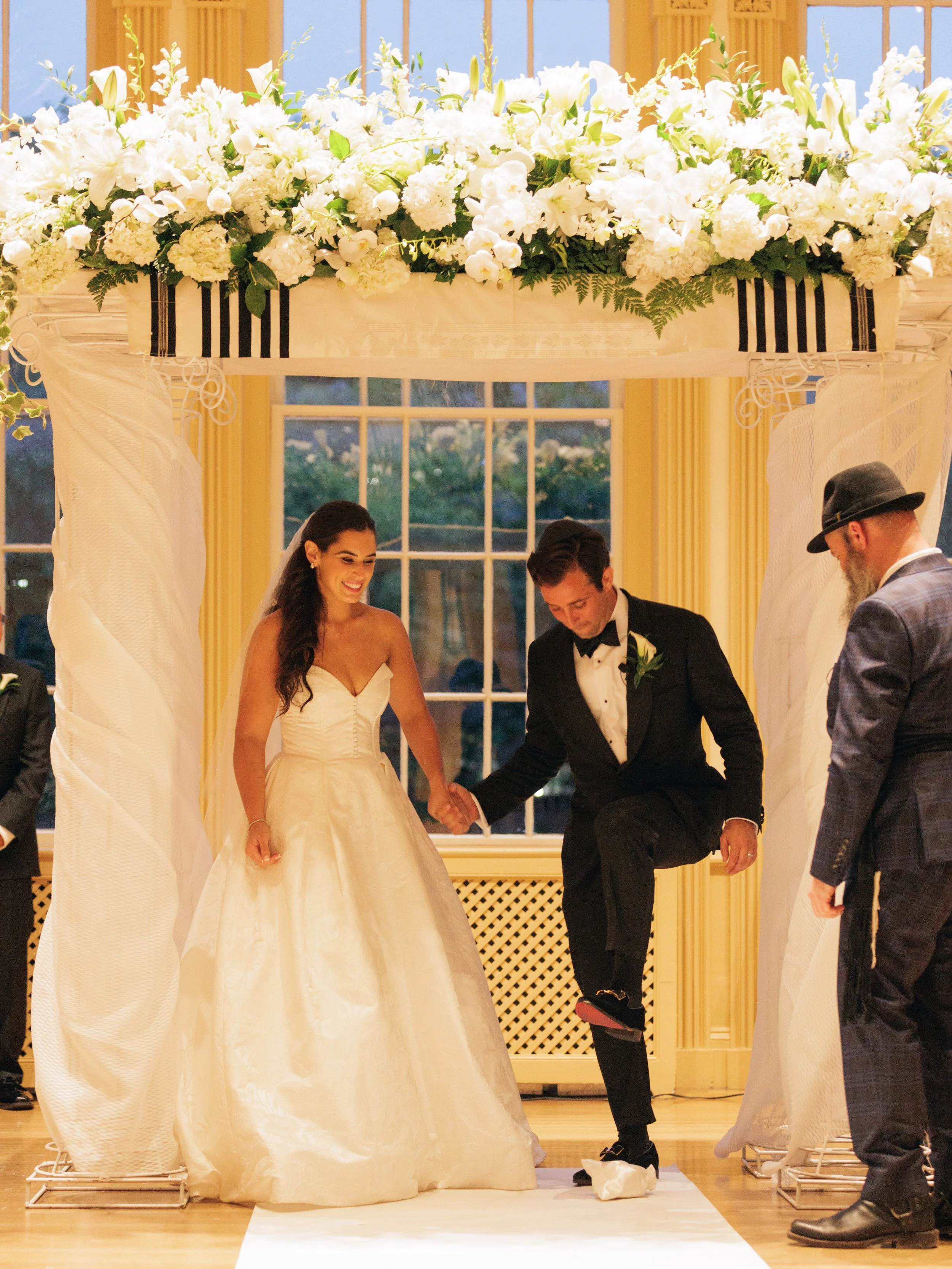 A wedding ceremony with the bride and groom holding hands, standing under a floral arch with white flowers and greenery, in a decorated room with large windows.