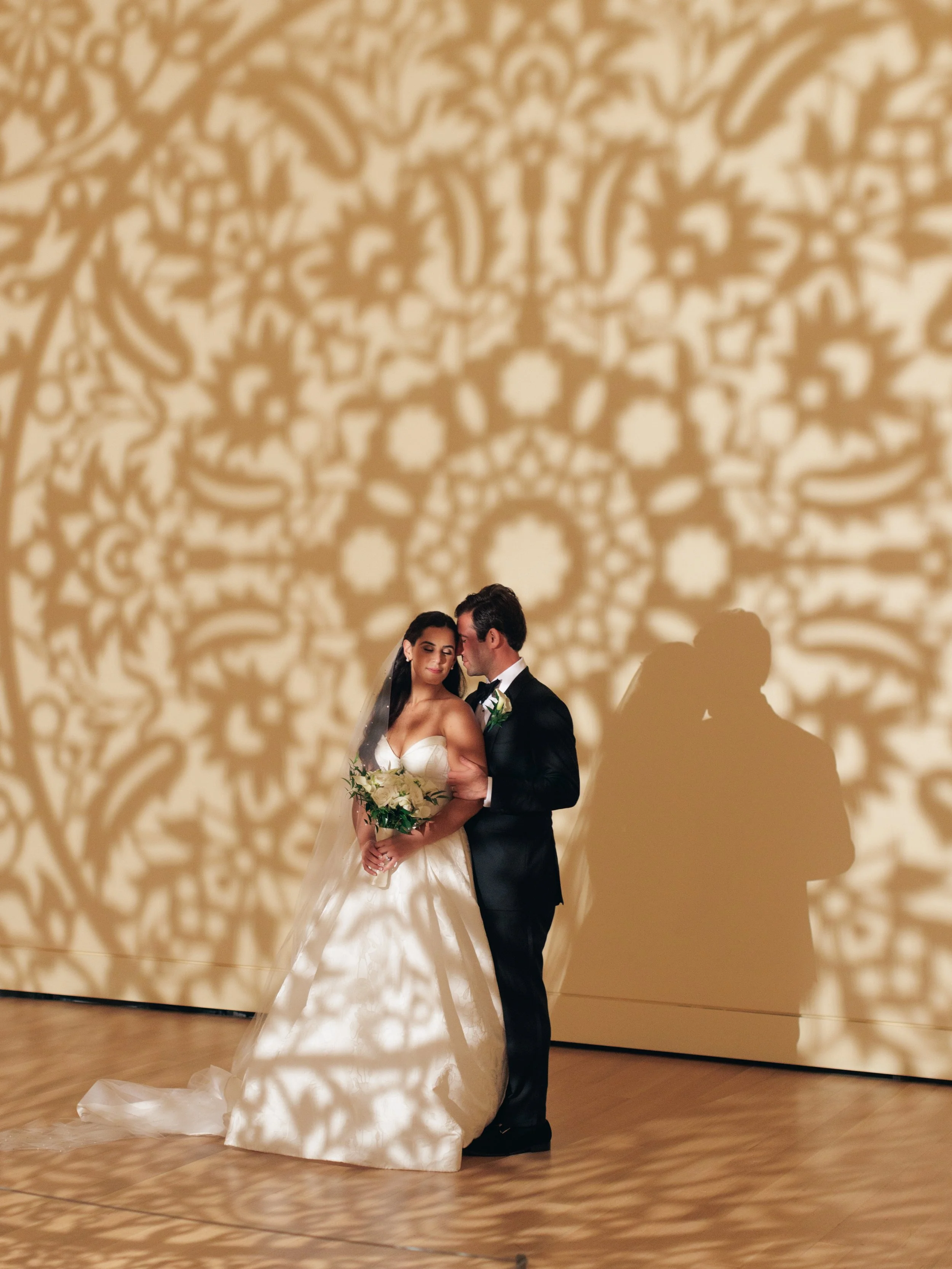 A bride and groom standing close together during their wedding reception, with intricate shadow patterns on the wall behind them.