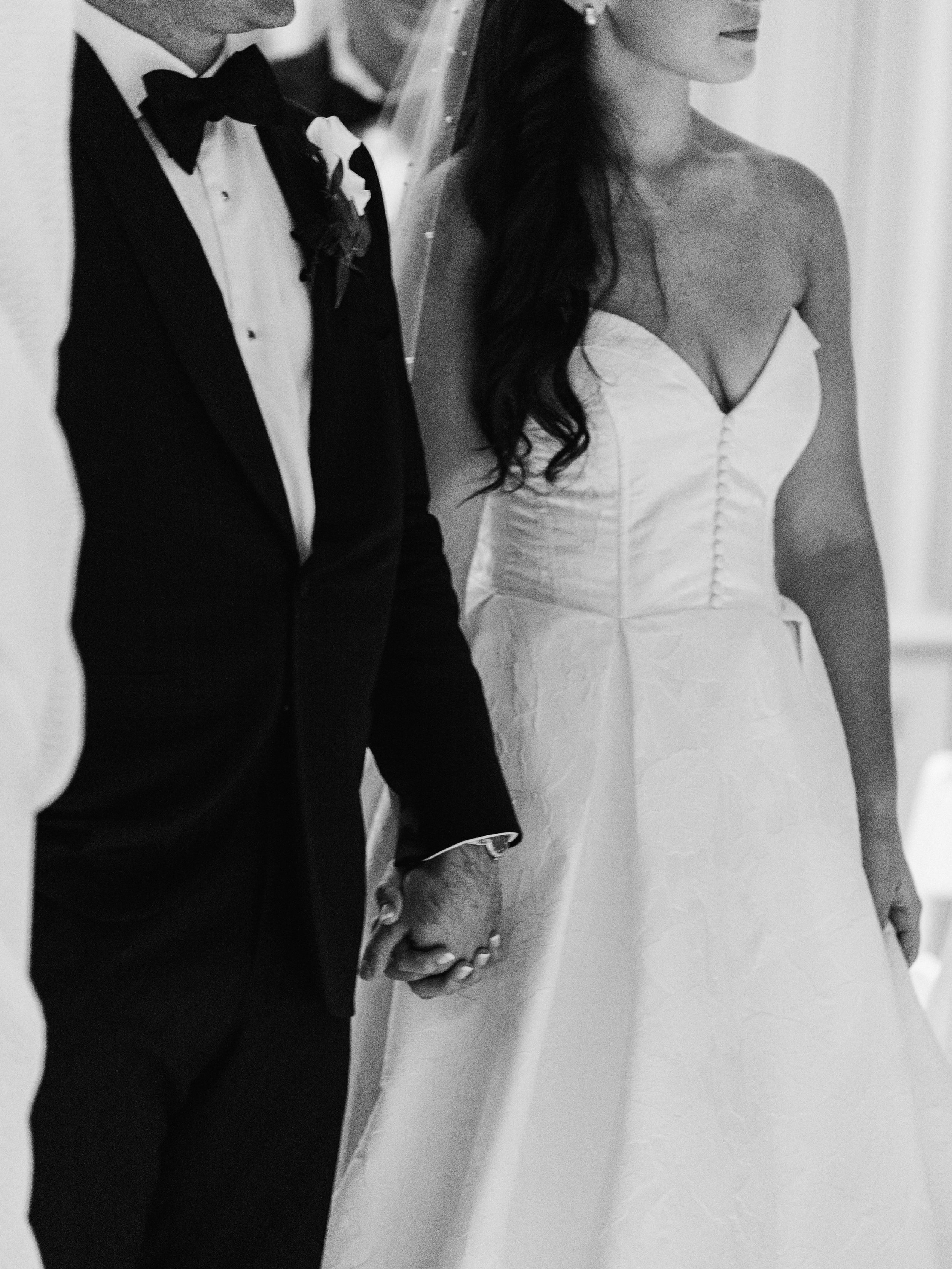 A close-up of a bride and groom holding hands during their wedding ceremony, both dressed in formal wedding attire, with the groom in a tuxedo and the bride in a strapless wedding gown.