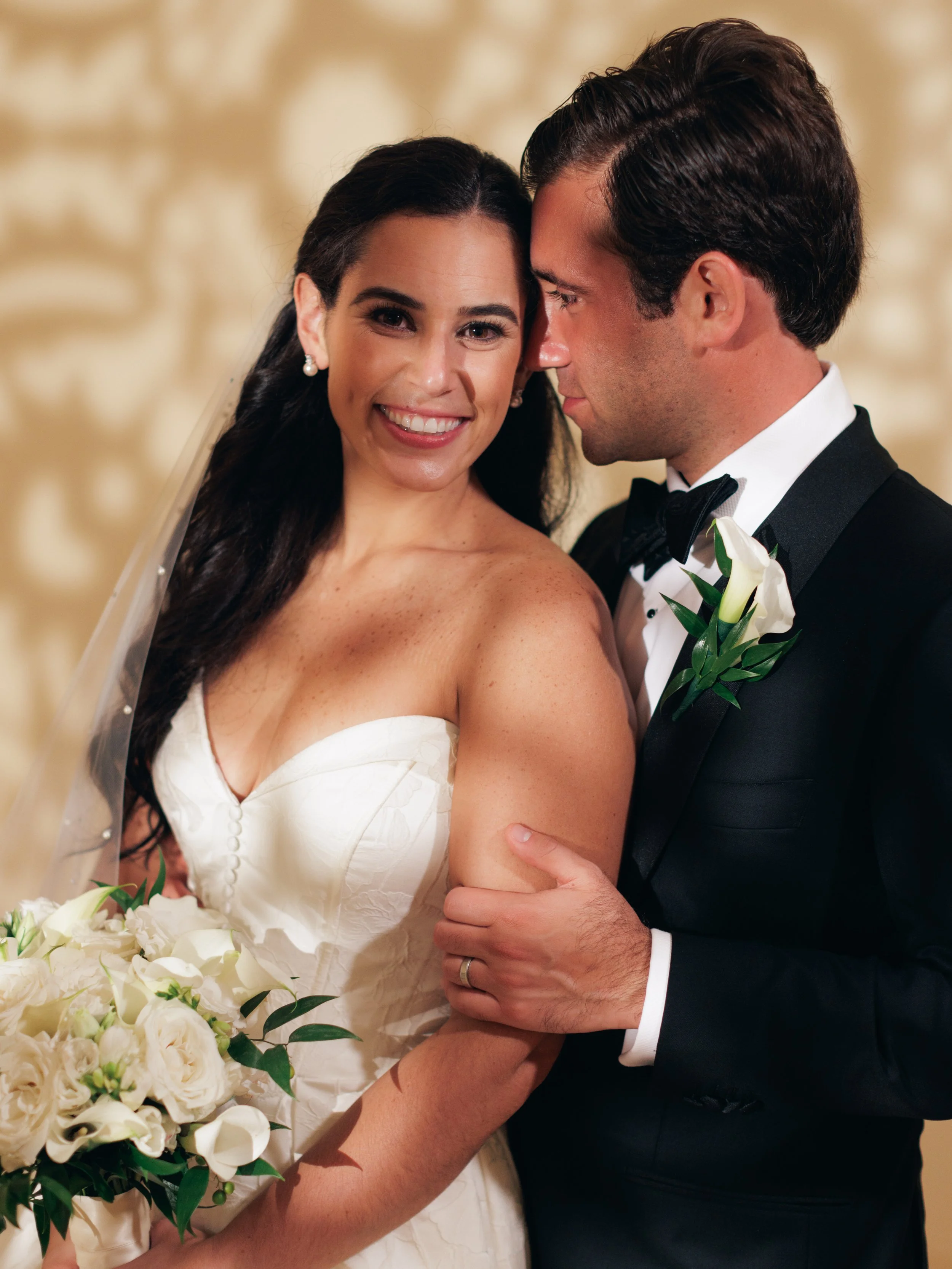 A bride and groom close together at their wedding, smiling, with the bride in a strapless white dress and the groom in a black tuxedo, holding hands.
