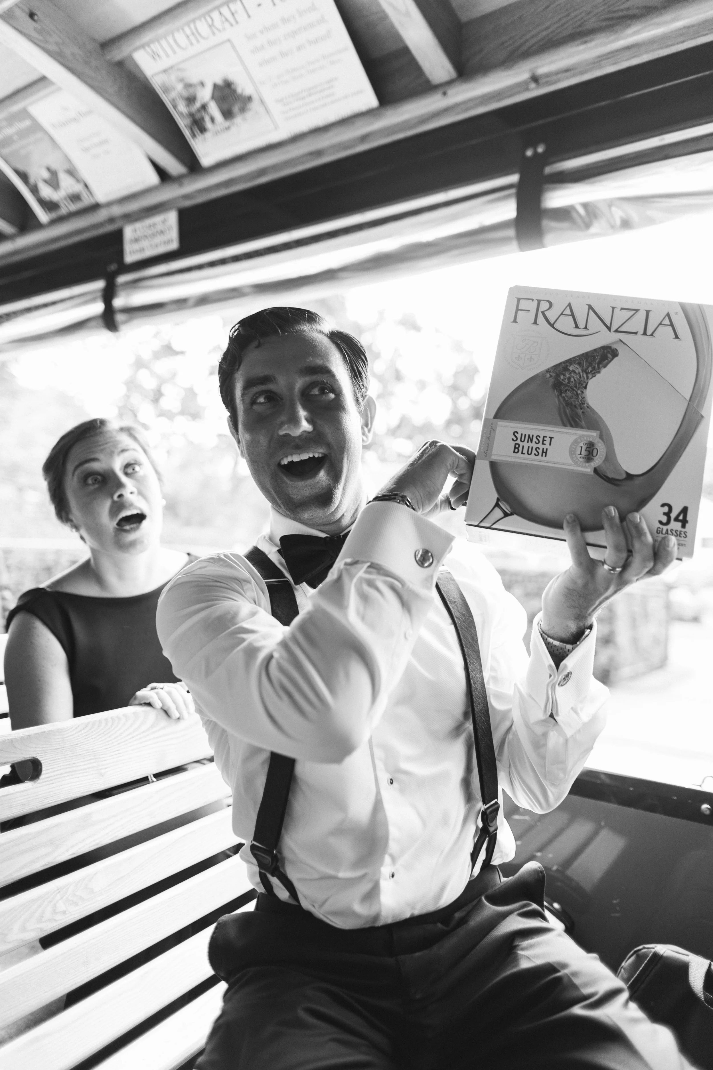Black and white photo of two surprised men sitting on a wooden bench, with one man holding a magazine titled 'Franzia Sunset Blush' and smiling at the camera, a woman sitting next to them with a shocked expression, in an outdoor setting with a wooden