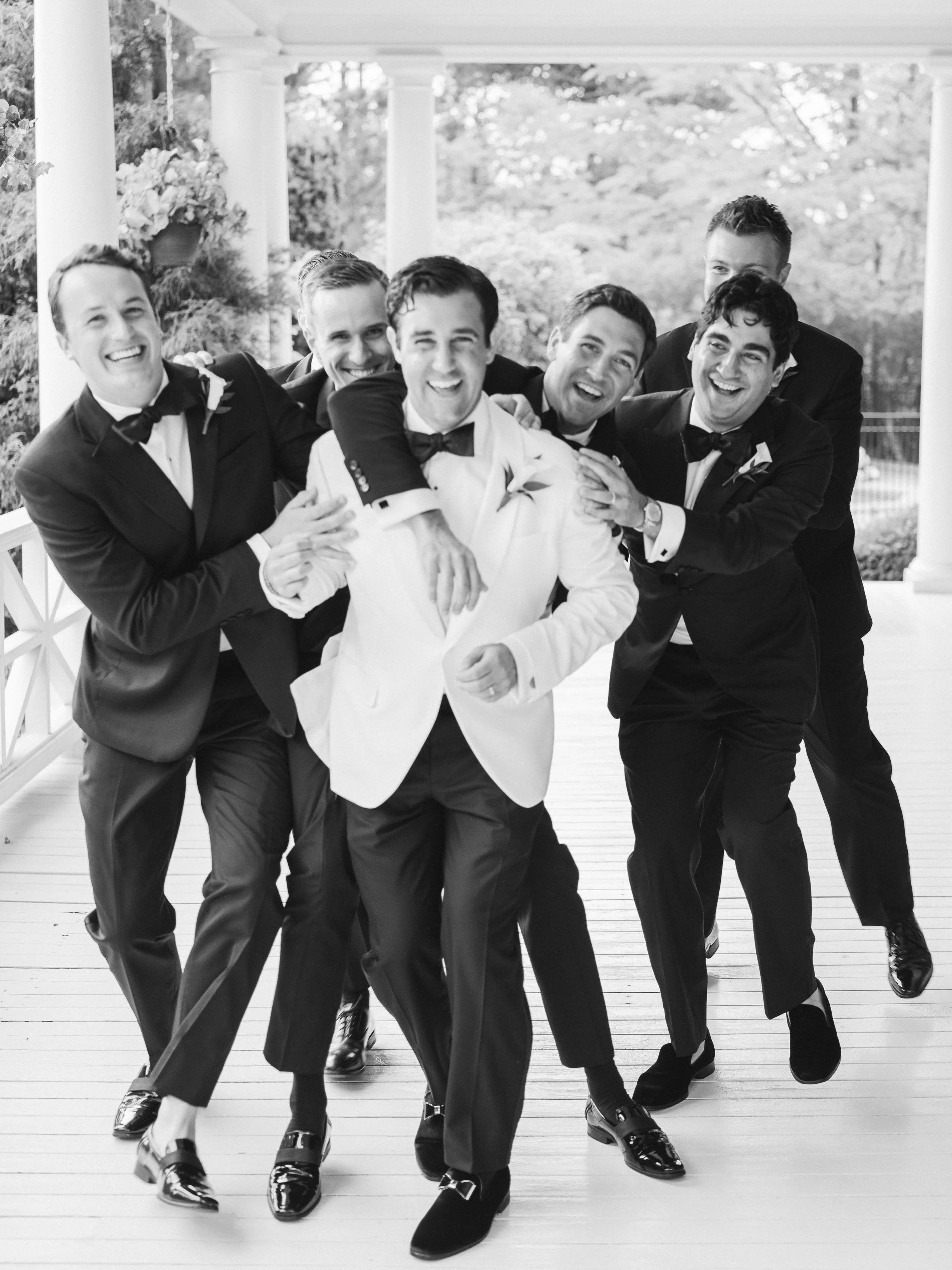 A group of seven men in tuxedos and bow ties are playfully posing together on a porch with columns, outdoors and surrounded by trees.