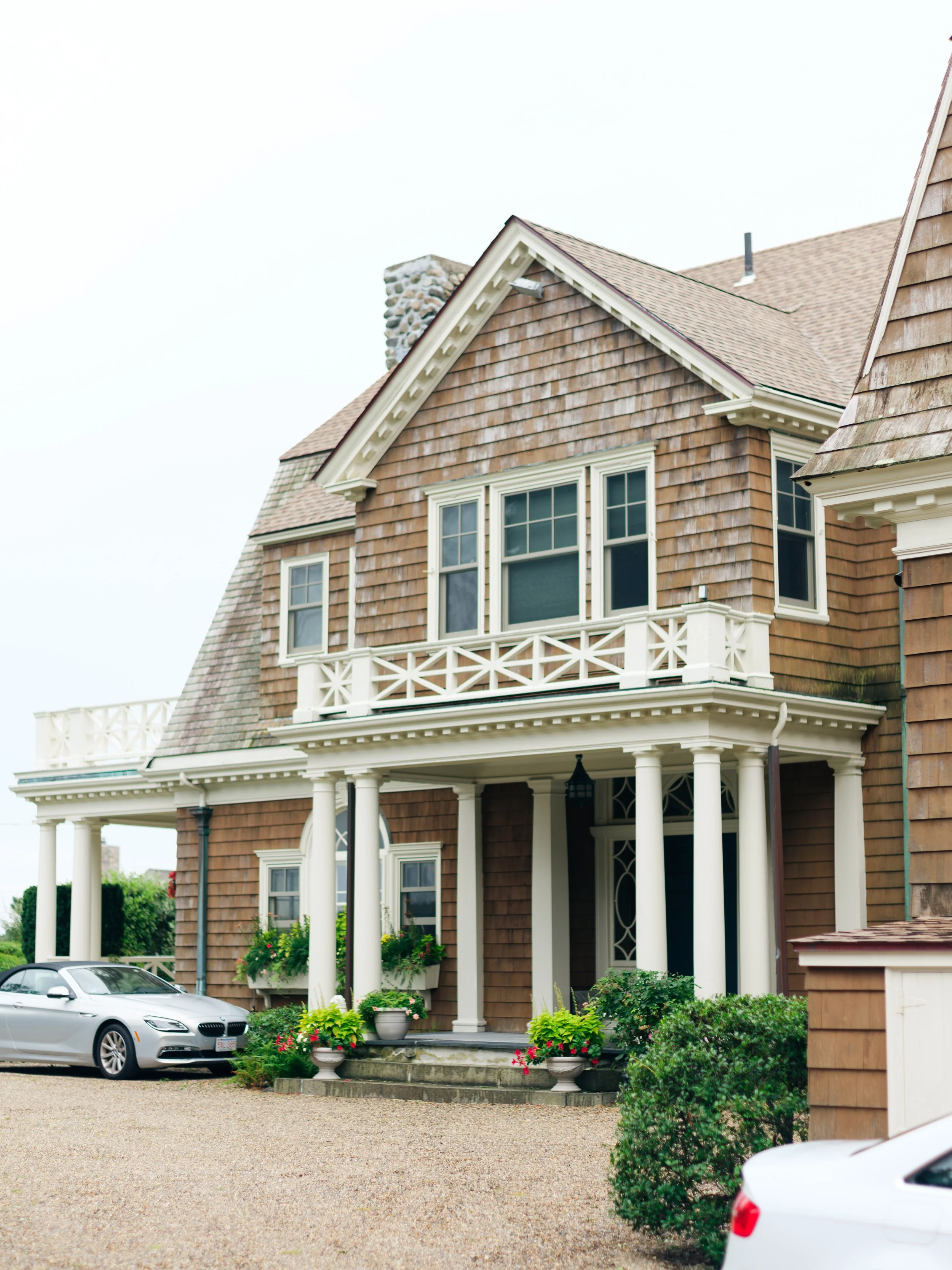 A large, two-story house with brown shingles, large white-framed windows, and a front porch with white columns. There are flower pots and greenery near the porch steps, and two parked cars are visible in the driveway. The sky is overcast.