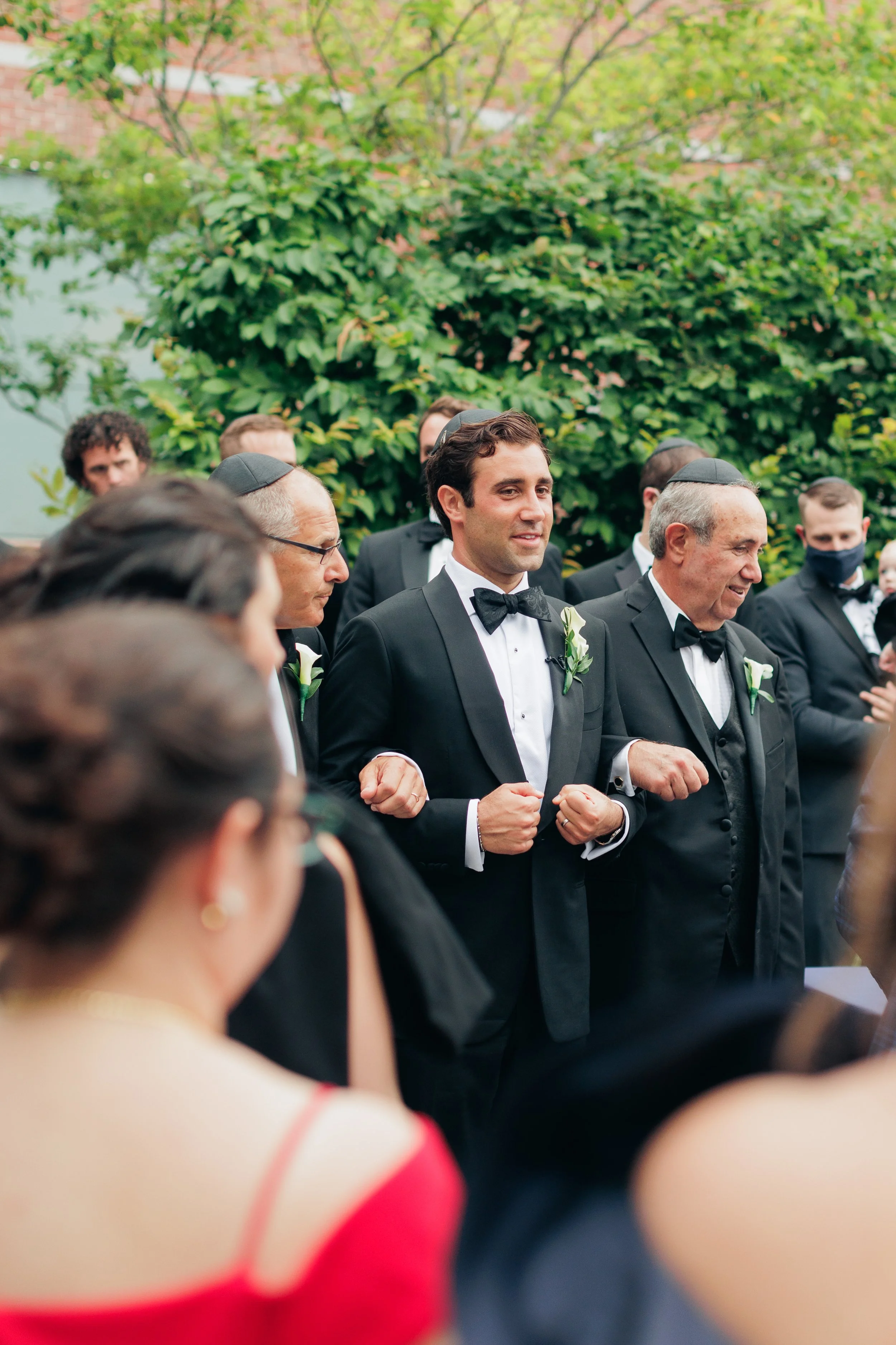Men in tuxedos and women dressed up in formal attire attending a wedding ceremony outdoors, with greenery and trees in the background.