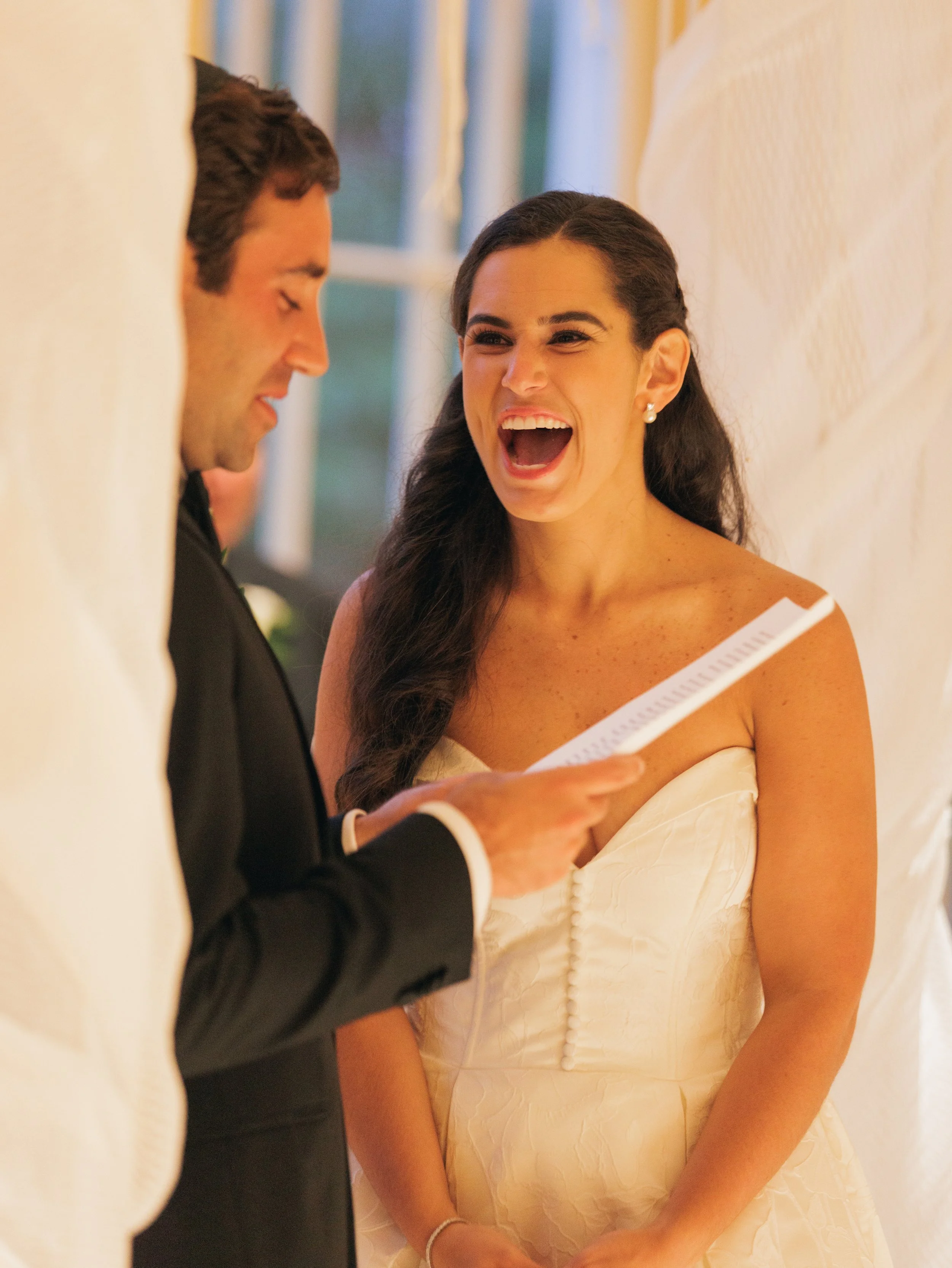 A couple getting married, the woman is wearing a white wedding dress and the man is in a black tuxedo. The woman is smiling with her mouth open while the man looks at her and holds a piece of paper.