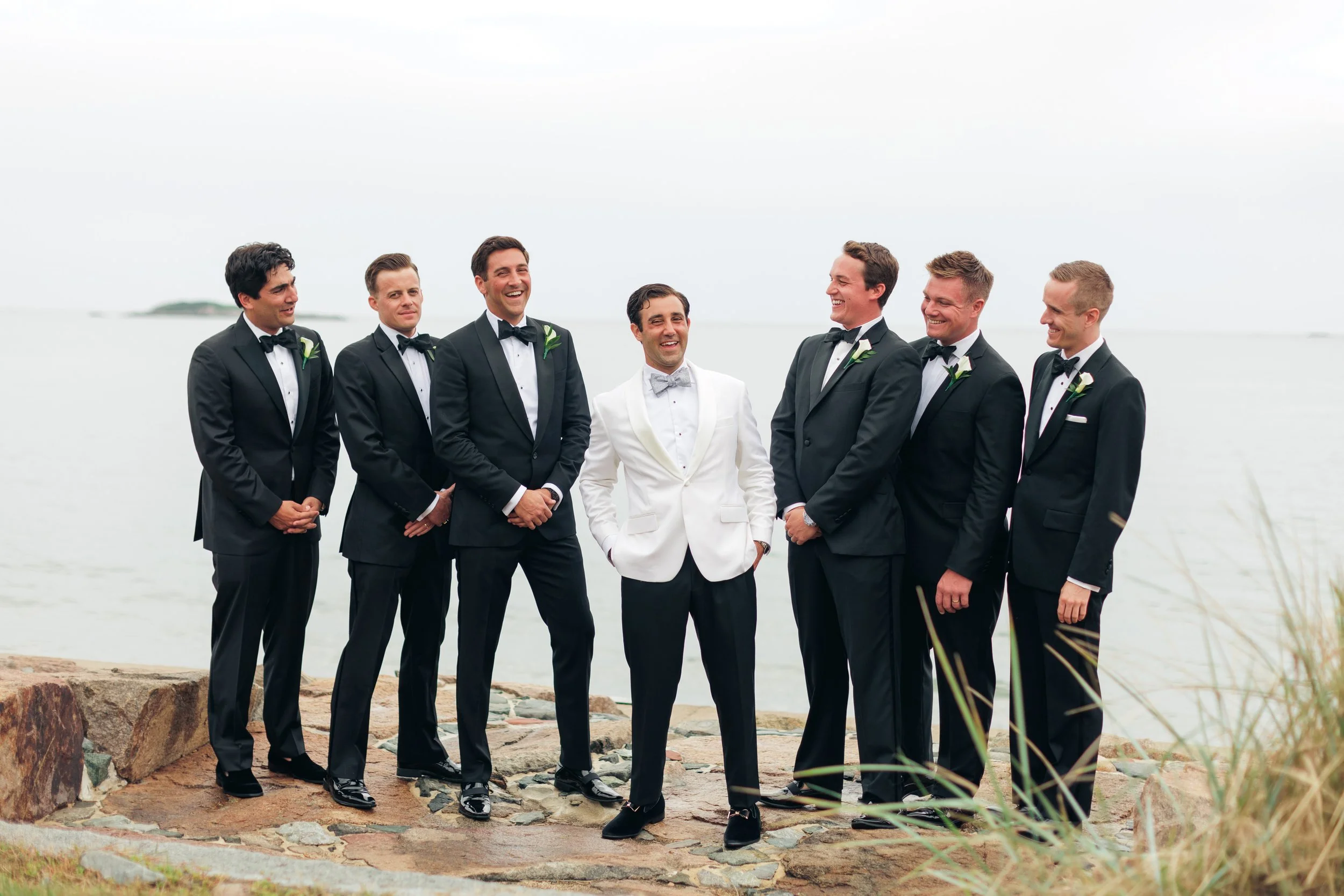 A group of seven men dressed in tuxedos, standing outdoors by the water, smiling and enjoying each other's company.