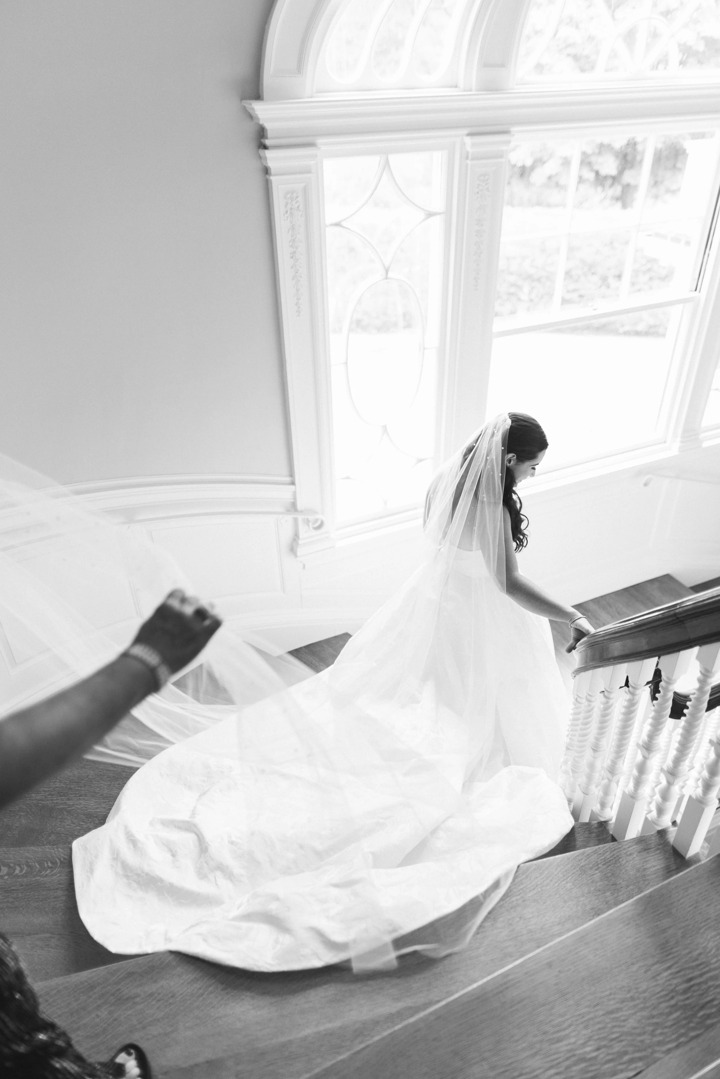 A bride with long dark hair and a veil, smiling, as she descends a staircase covered with a white cloth. An arm with a watch is visible, holding the cloth.