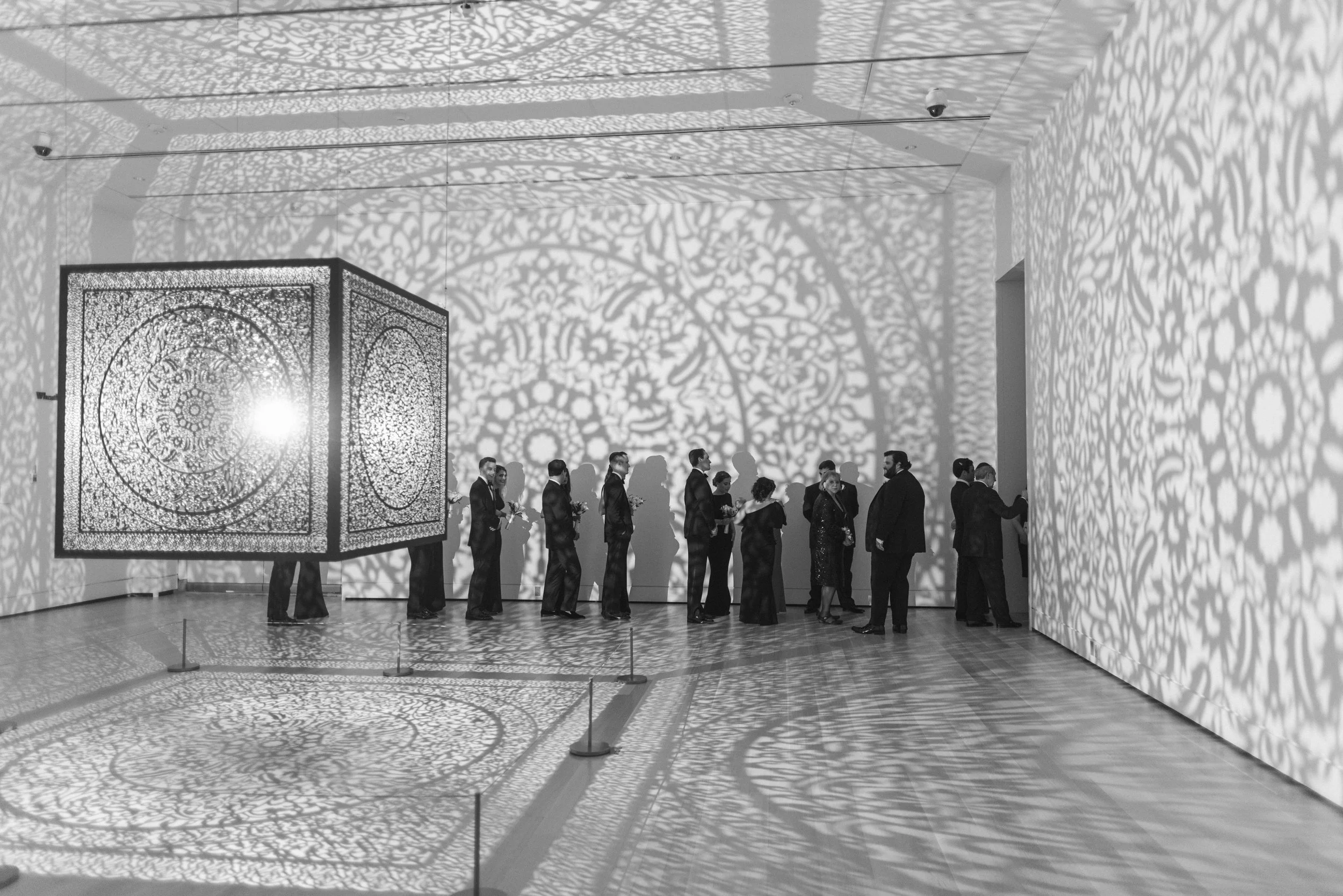 Group of people gathered in an art gallery or exhibition space, with shadows of intricate patterns cast on the walls and floor, and a large decorative box-like structure with detailed filigree patterns.