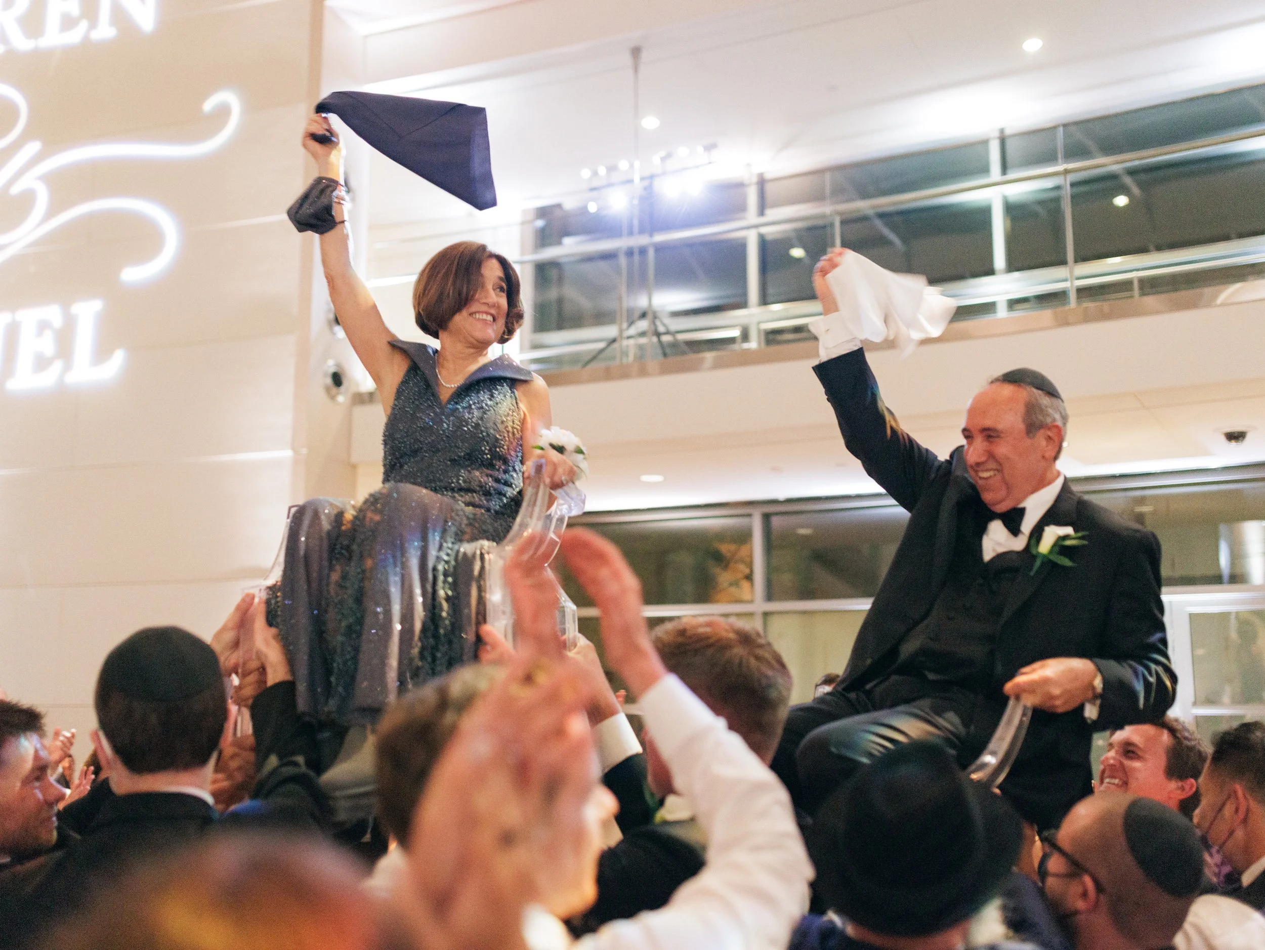 A woman in a sleeveless dress and a pearl necklace is being lifted in the air by a crowd at a formal event. A man in a suit is sitting nearby, smiling and holding a tissue, as they celebrate together.