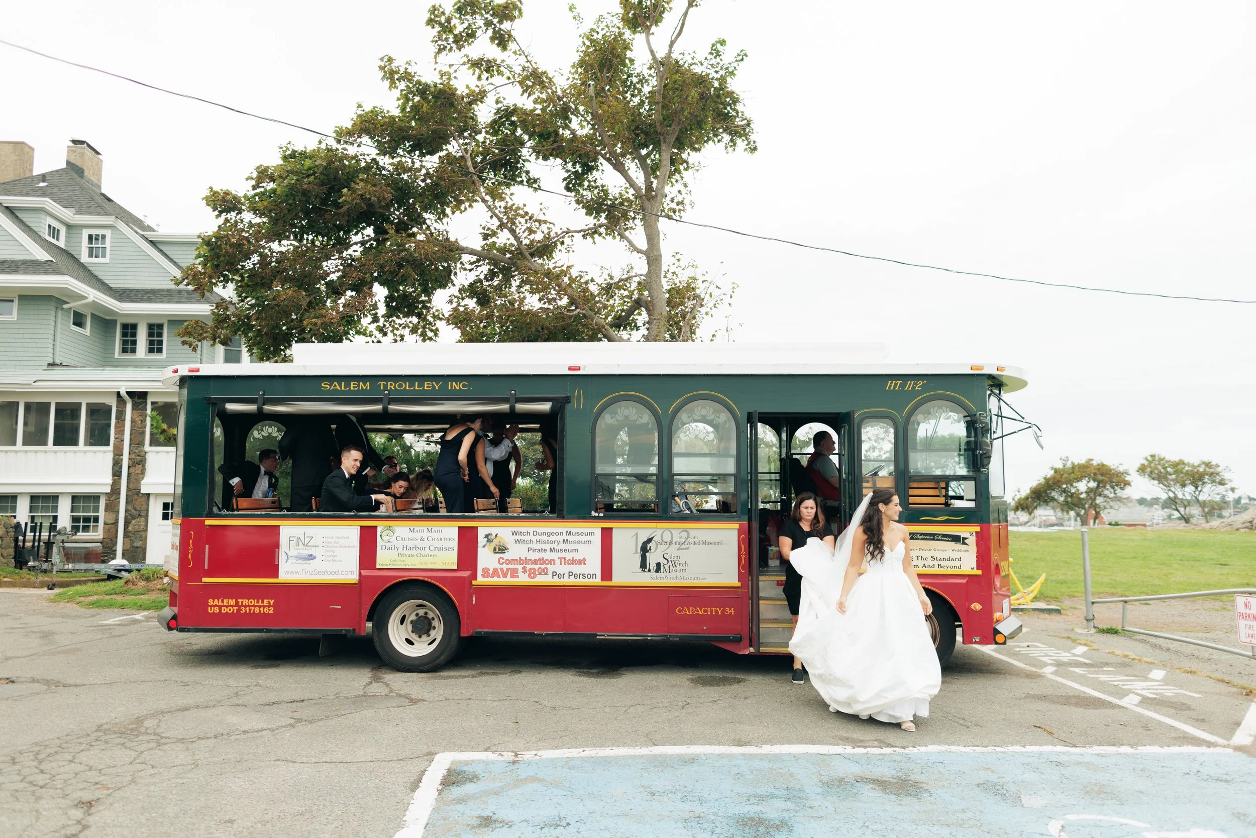 A red trolley with a green top, labeled 'Salem Trolley Inc.', is parked outdoors near a grassy area. People, some dressed in wedding attire, are sitting and standing around the trolley. A woman in a white wedding dress and veil is standing beside two